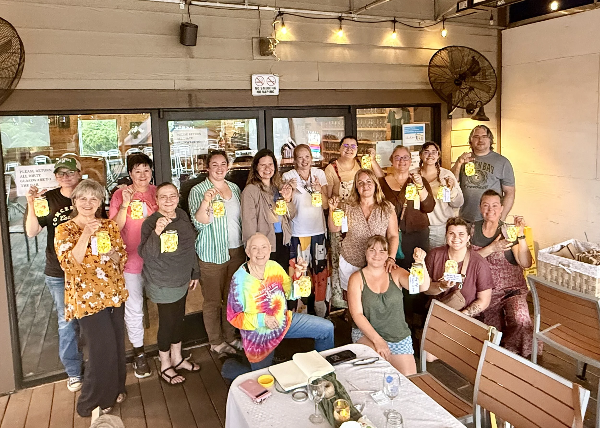 A group of people holding lanterns gathered inside a restaurant or cafe, smiling and posing for the photo, with string lights and fans on the walls.