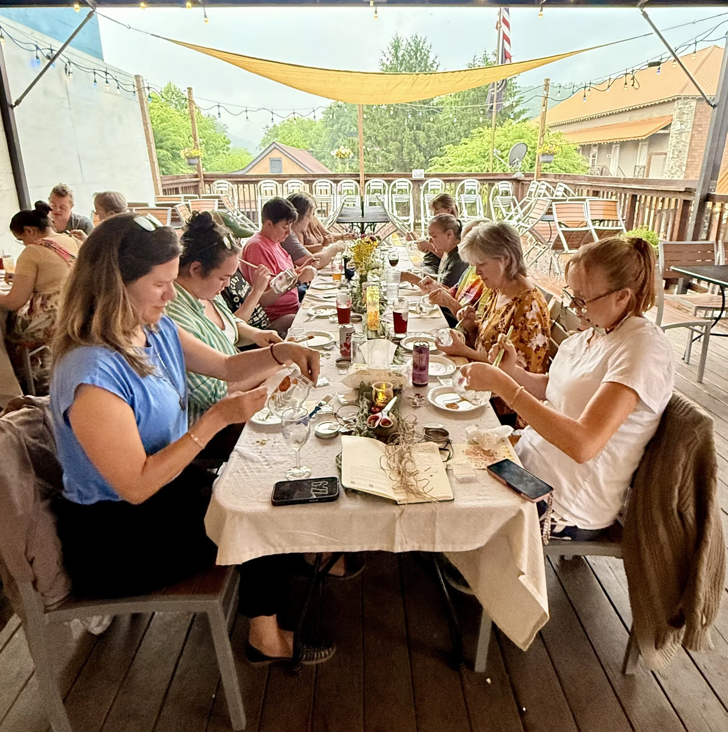 People sitting at a long dining table on an outdoor patio, with string lights and trees in the background, enjoying a meal together.