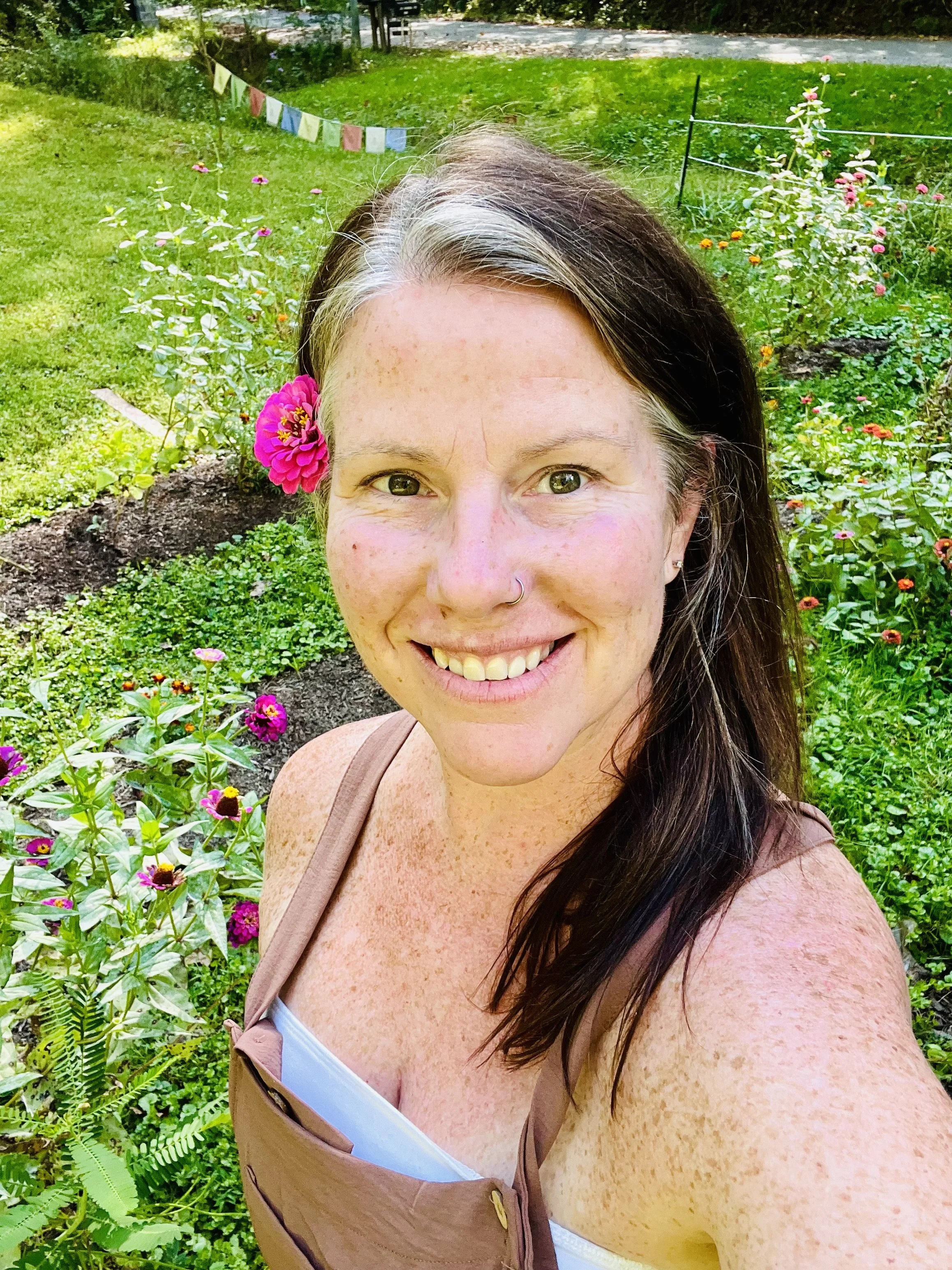 A smiling woman with dark hair, freckles, a nose ring, and pink flower in her hair, taking a selfie in a garden with colorful flowers and green grass.