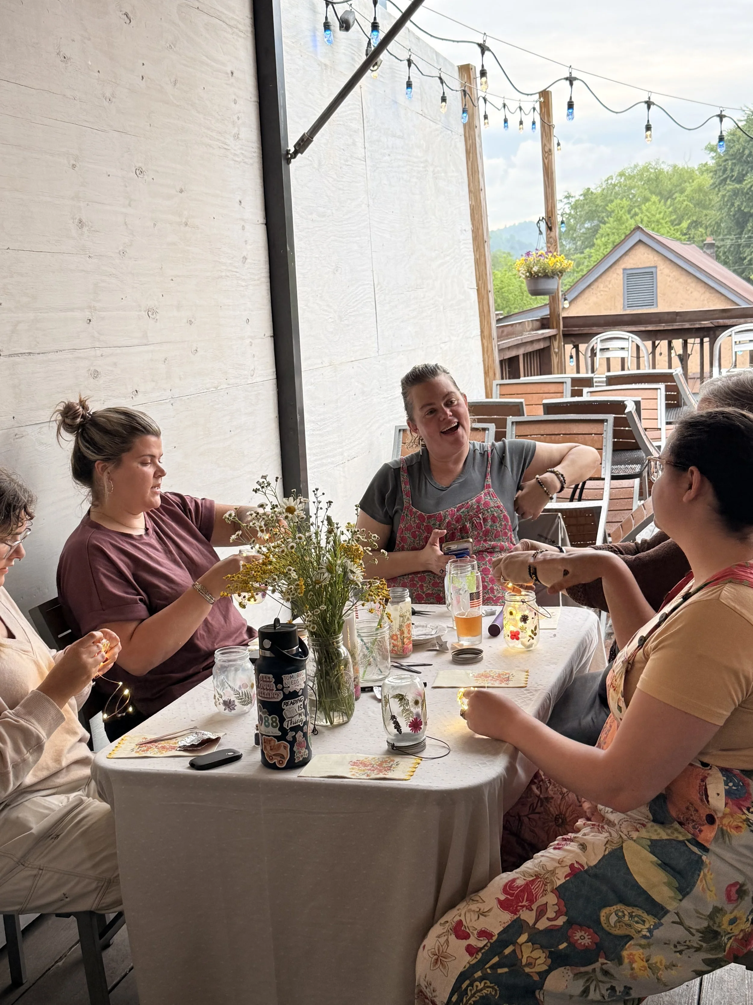 A group of people sitting around a dining table on a porch, enjoying each other's company. There are string lights hanging above, a vase with flowers, and drinks on the table. A woman in the center is smiling and talking, wearing a gray t-shirt and f