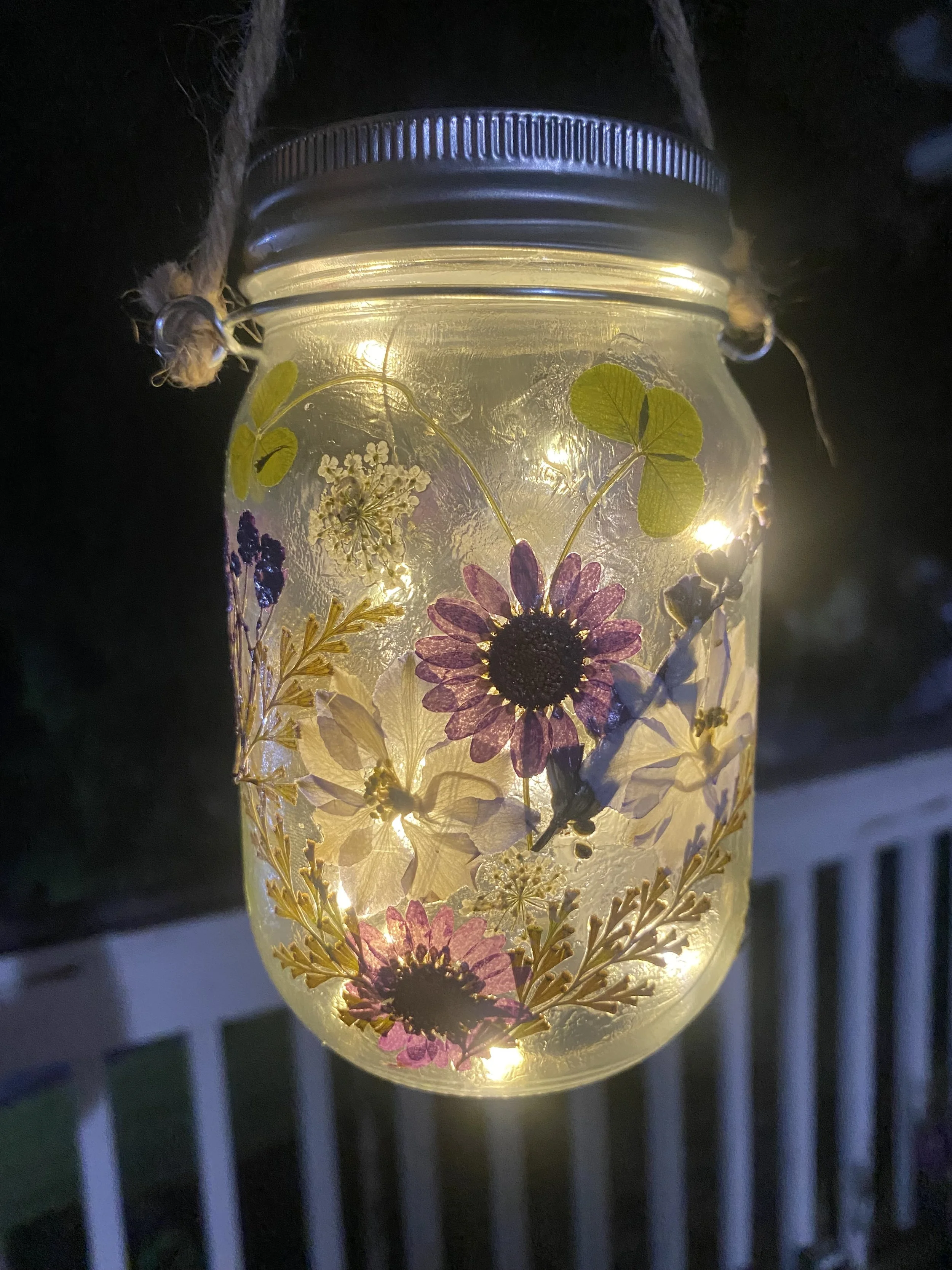 A decorative glass jar with pressed flowers and leaves inside, illuminated by a warm light, hanging on a rope outside at night.