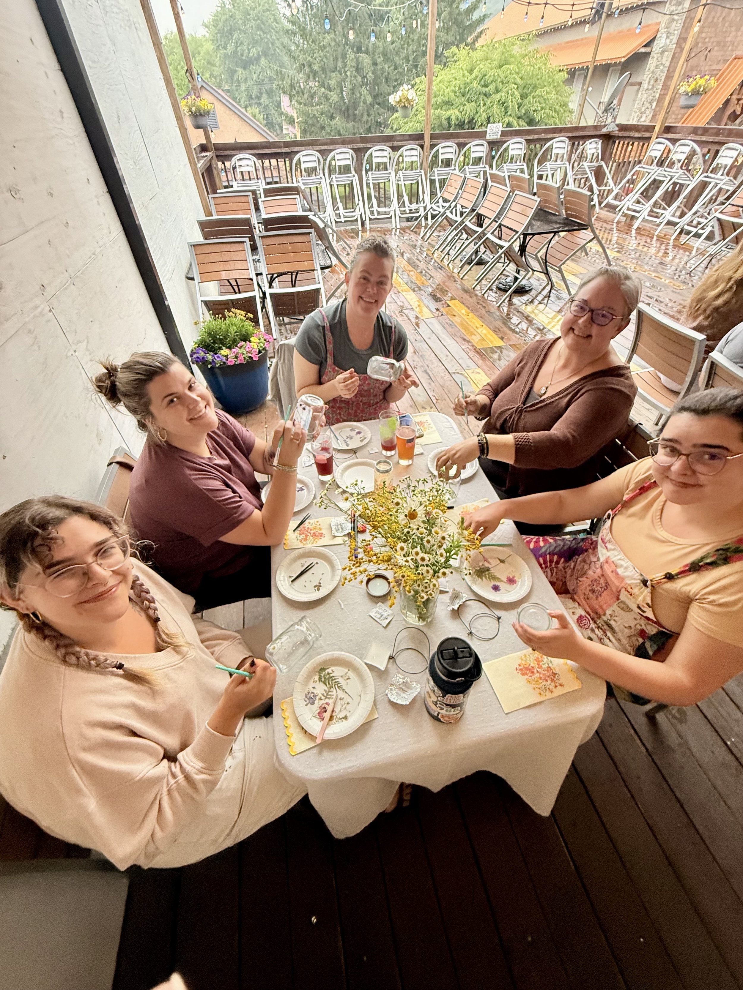 Group of women sitting around a table outdoors, celebrating with drinks and flowers during a rainy day.