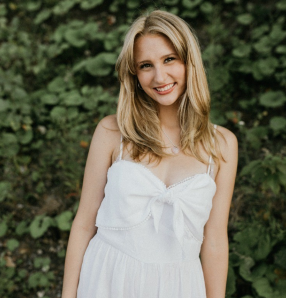A young woman with blonde hair smiling in front of green foliage, wearing a white sleeveless dress.