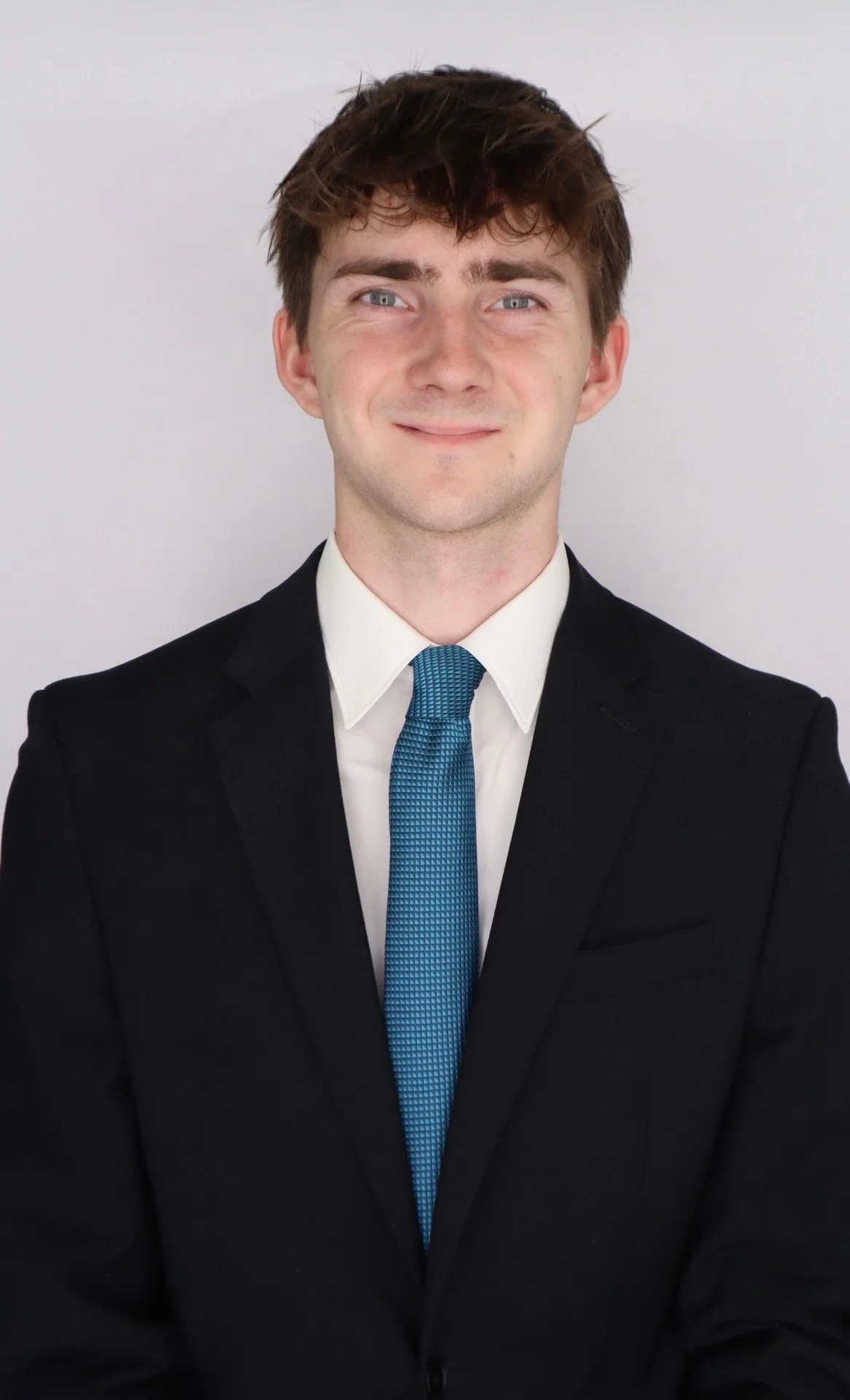 Young man in formal black suit with white shirt and blue tie, smiling against a plain light gray background.
