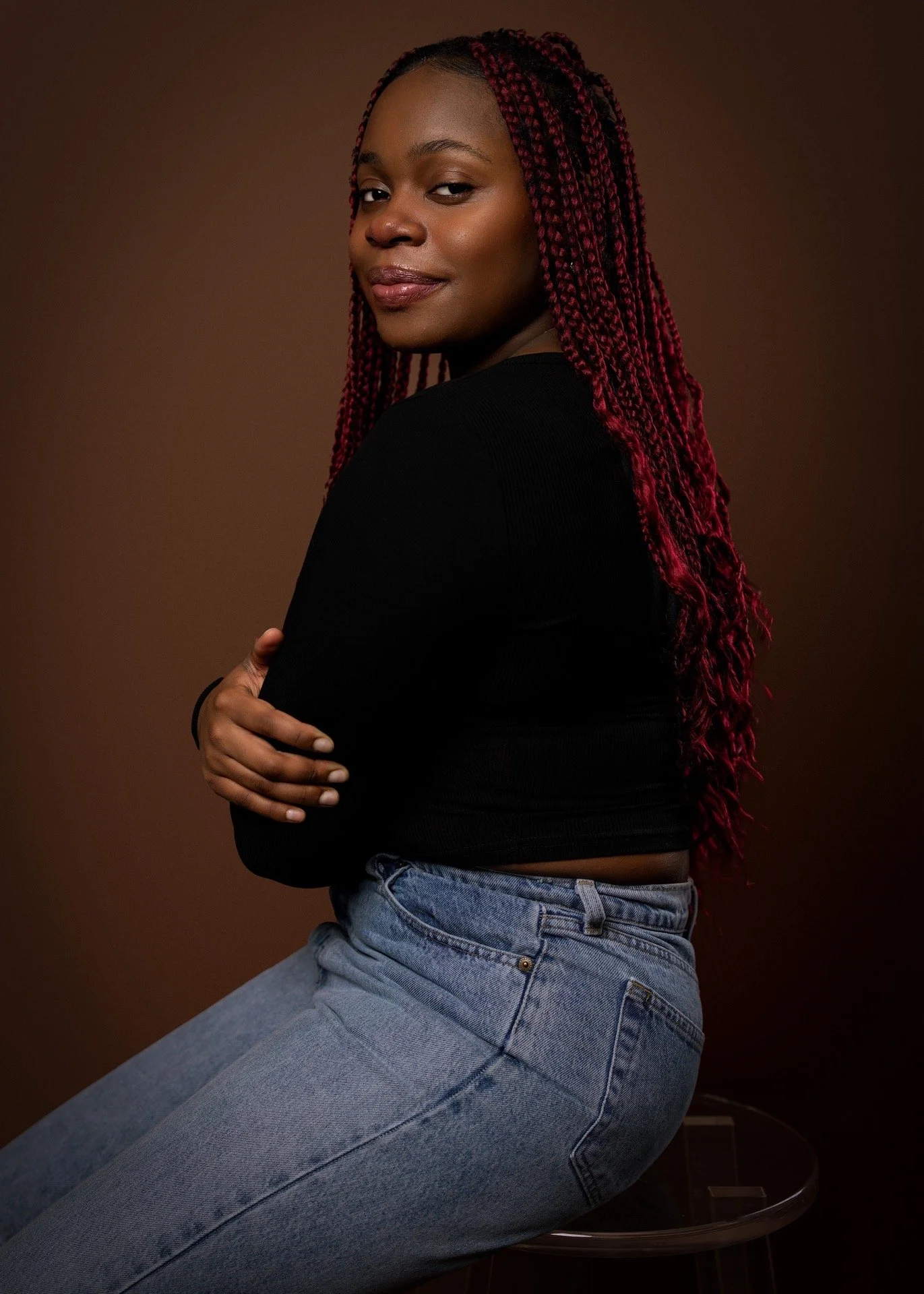 A young woman with red braided hair, wearing a black long-sleeve shirt and jeans, sitting intently on a stool against a dark brown background.