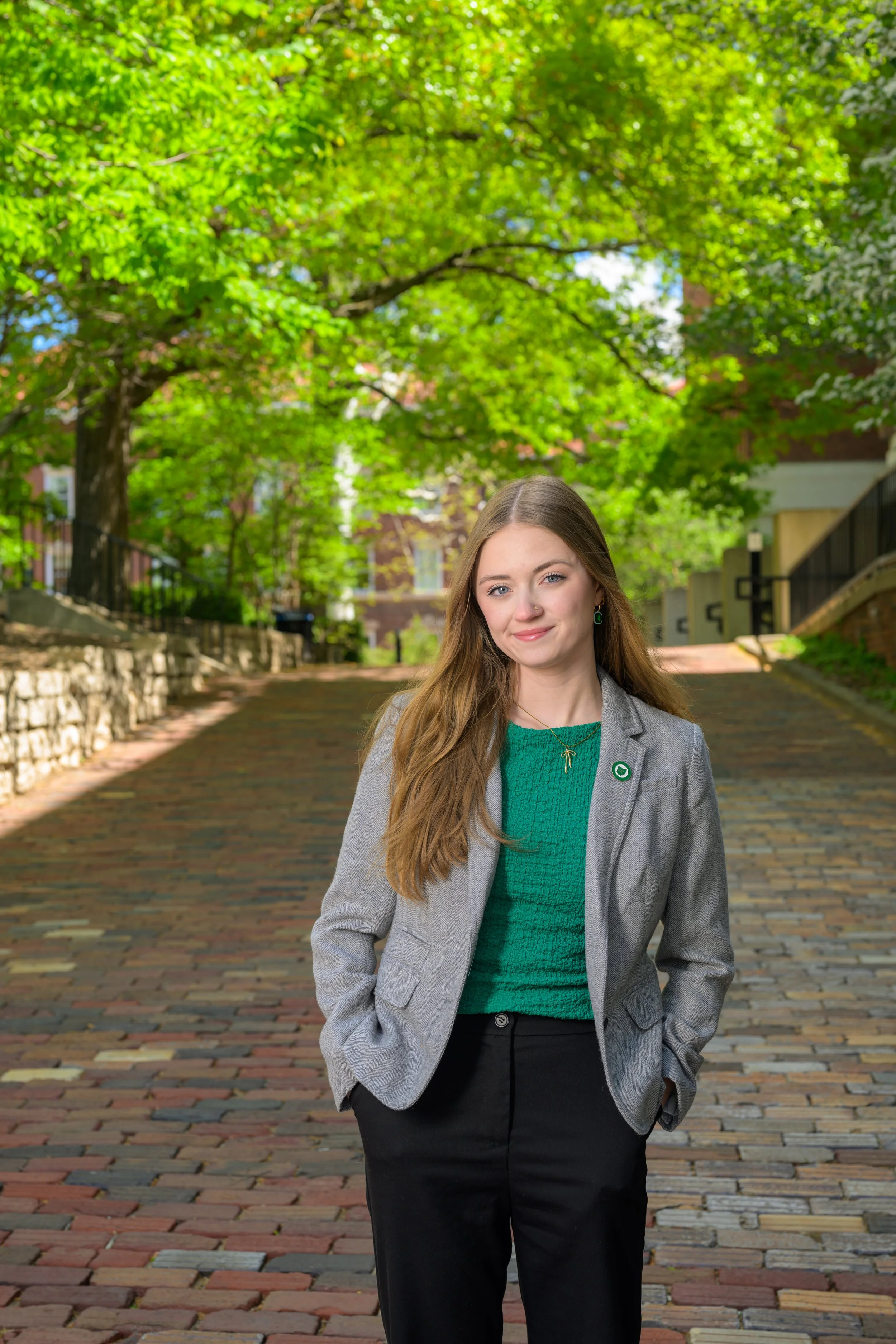 A woman with long hair, wearing a gray blazer, green sweater, and black pants, standing outdoors on a brick path with green trees and buildings in the background.