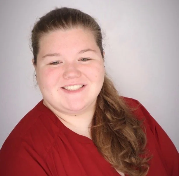 A young woman with long brown hair in a side braid, wearing a red top, smiling against a plain light gray background.