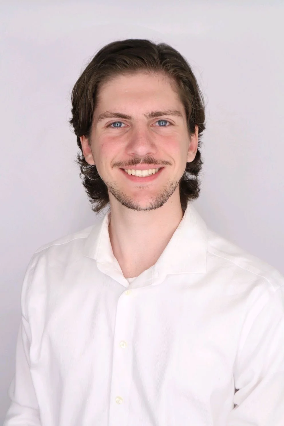A young man with brown wavy hair, blue eyes, and a light beard, smiling and wearing a white button-up shirt. Portrait against a plain, light-colored background.