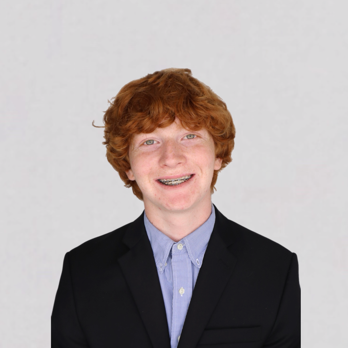 Young red-haired teenage boy with braces, wearing a black blazer and a light blue button-up shirt, smiling against a plain light gray background.