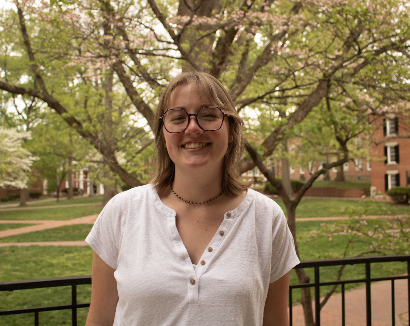 Smiling young woman with glasses and a white shirt standing outdoors near a black railing, with blossoming trees and brick buildings in the background.