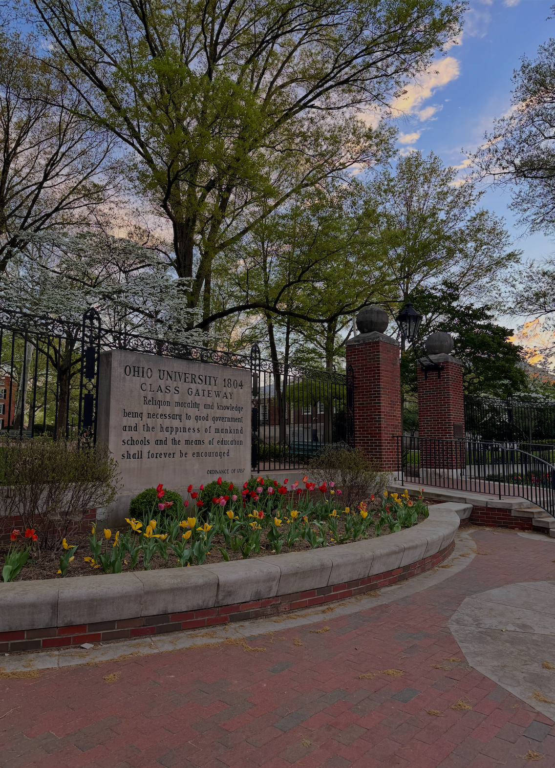 A stone sign at Ohio University with a quote, surrounded by blooming tulips and a brick pathway, with trees and a gated entrance in the background during sunset.