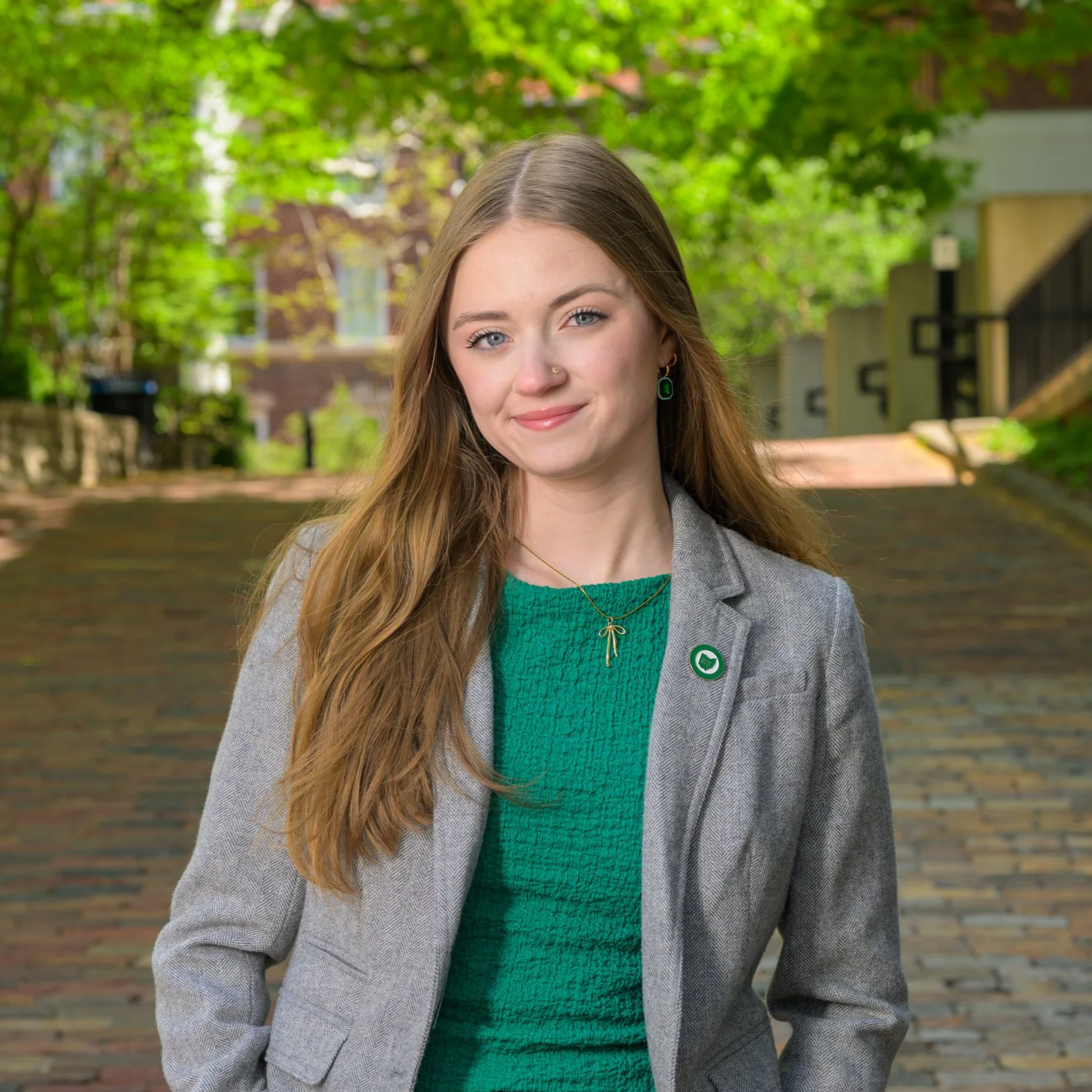 A young woman with long brown hair, wearing a grey blazer, green top, and jewelry, standing outside with green trees and buildings in the background.