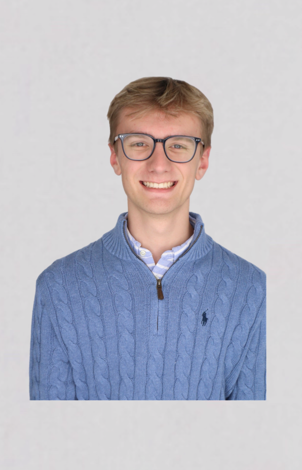 Portrait of a young man with glasses, smiling, wearing a blue cable-knit sweater and a collared shirt, standing against a plain background.