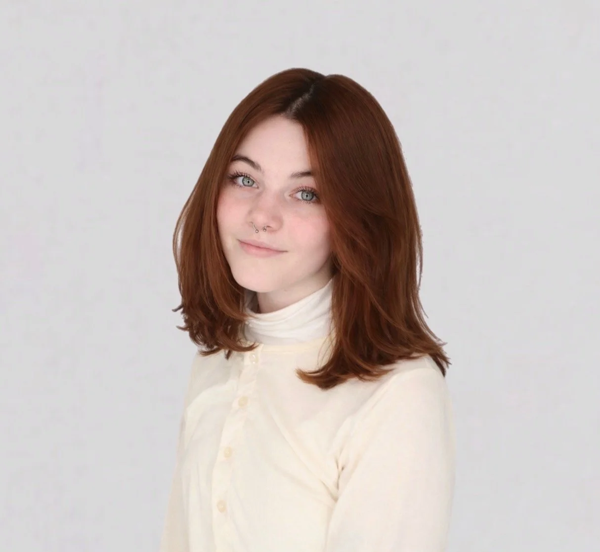 A young woman with red hair and light skin, wearing a cream-colored turtleneck, looking at the camera with a slight smile against a plain light gray background.