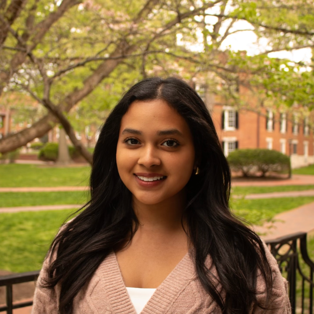 Young woman with long dark hair smiling outdoors on a campus green with trees and brick buildings in the background.