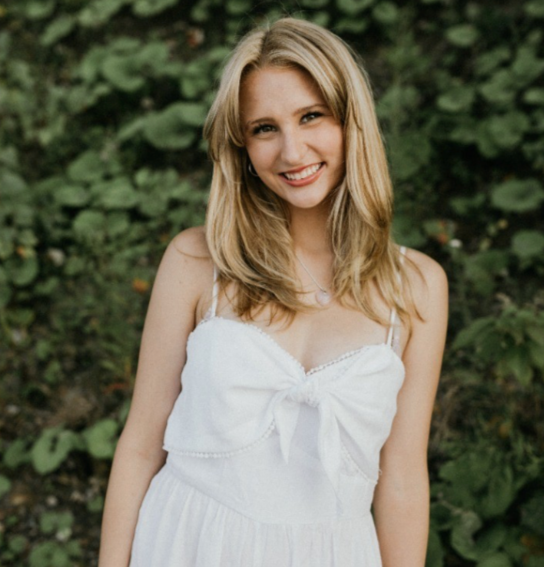 Young woman with blonde hair smiling outdoors in a white dress.