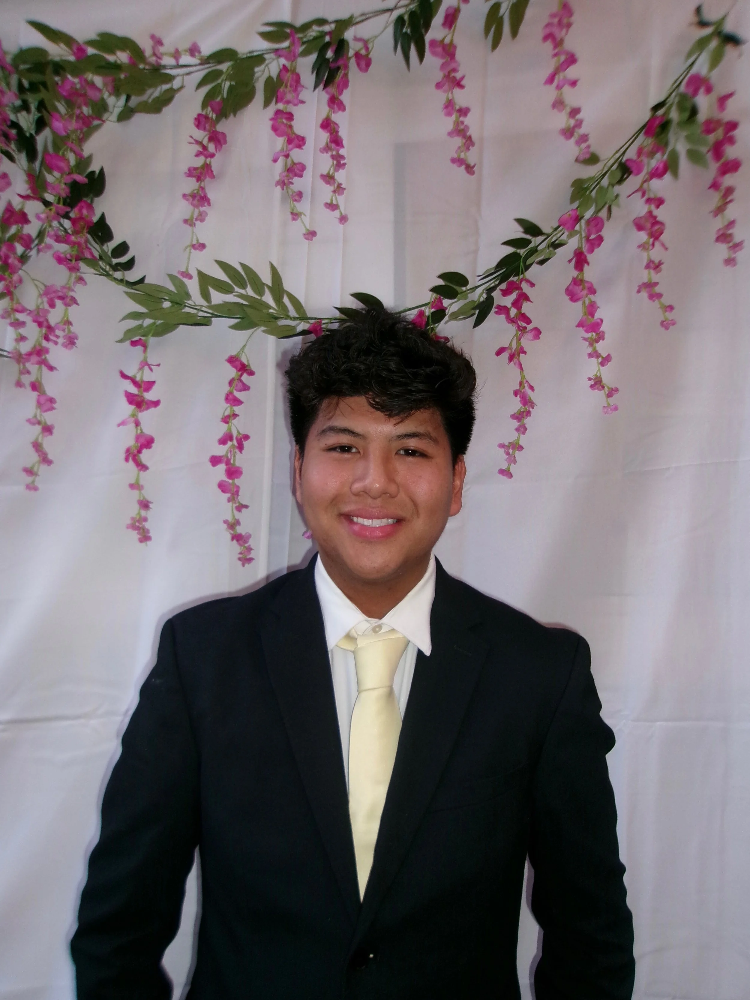 Young man in a black suit and cream-colored tie taking a selfie in front of a white backdrop with hanging pink flowers.