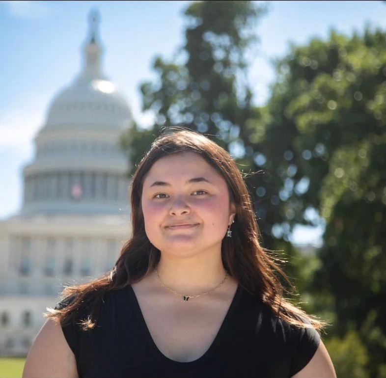 Young woman with long brown hair smiling outdoors with the U.S. Capitol in the background.