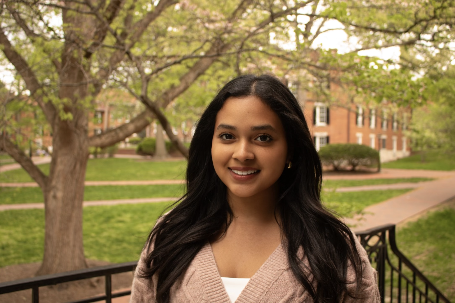 A smiling woman with long dark hair stands outdoors near a park bench, with green grass, trees, and a brick building in the background.