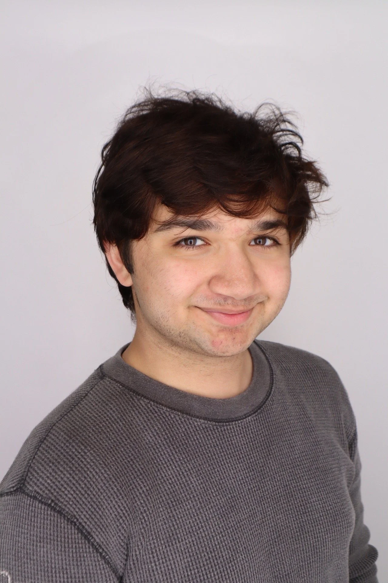 A young man with dark brown, tousled hair and light skin, smiling slightly, wearing a dark gray textured crewneck sweatshirt, standing against a plain gray background.