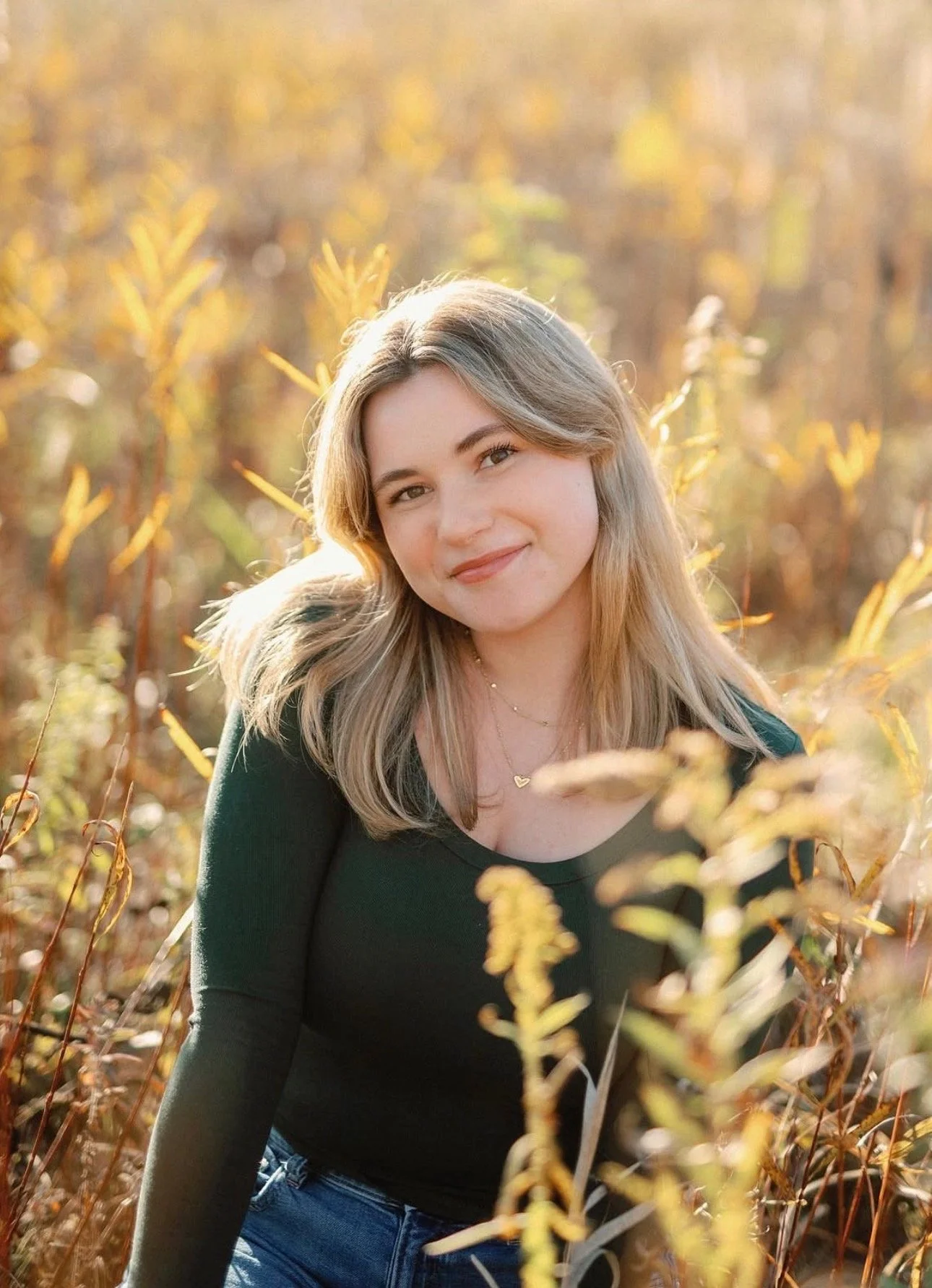 A young woman with shoulder-length blonde hair smiling outdoors in a field of yellow plants during sunset.