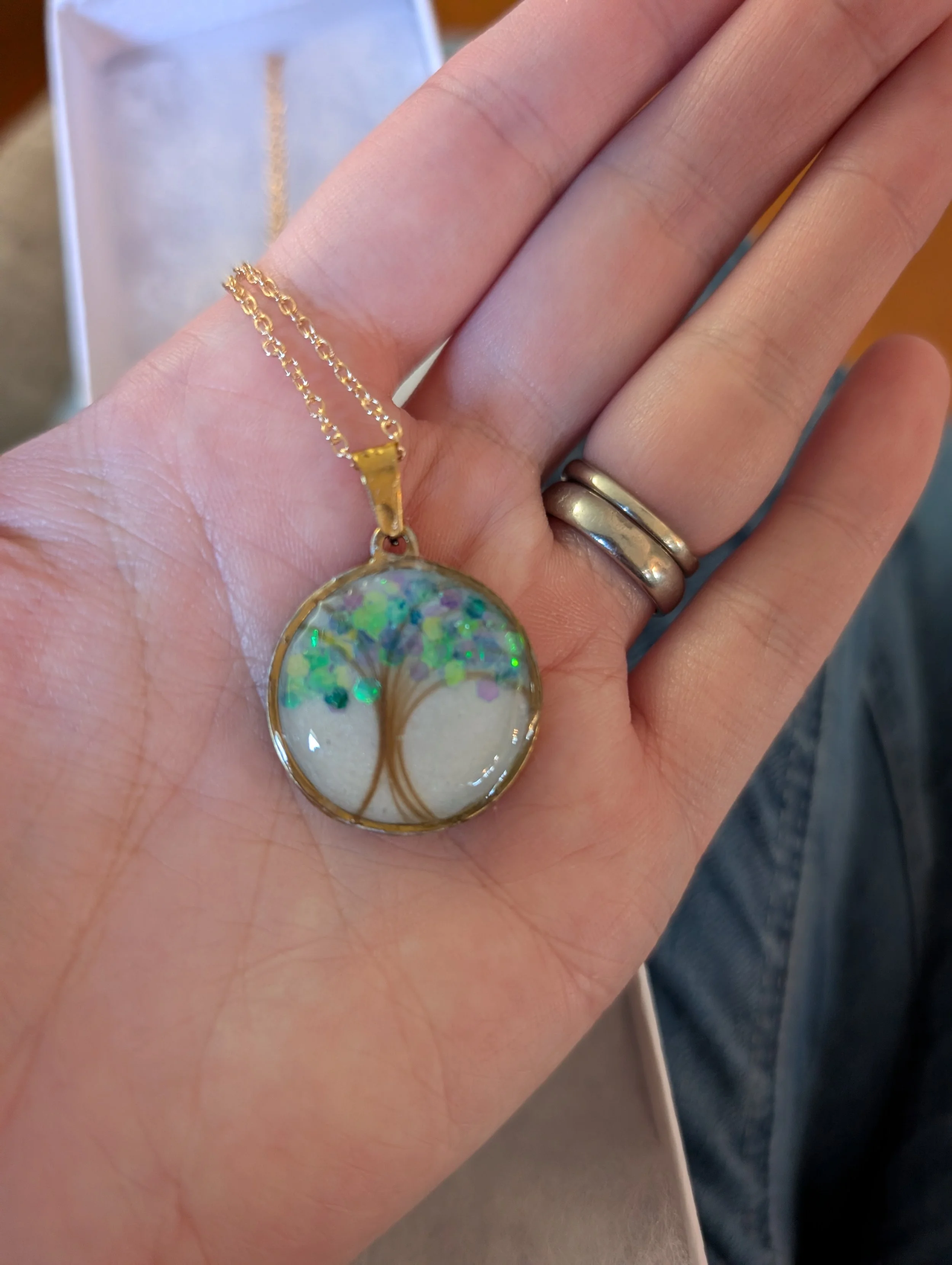 Close-up of a hand with a gold chain necklace and two rings, one gold and one silver, on the ring finger. The necklace has a circular pendant with iridescent colors and is shown over the palm of the hand. In the background, there is a blurred jewelry box or display case.