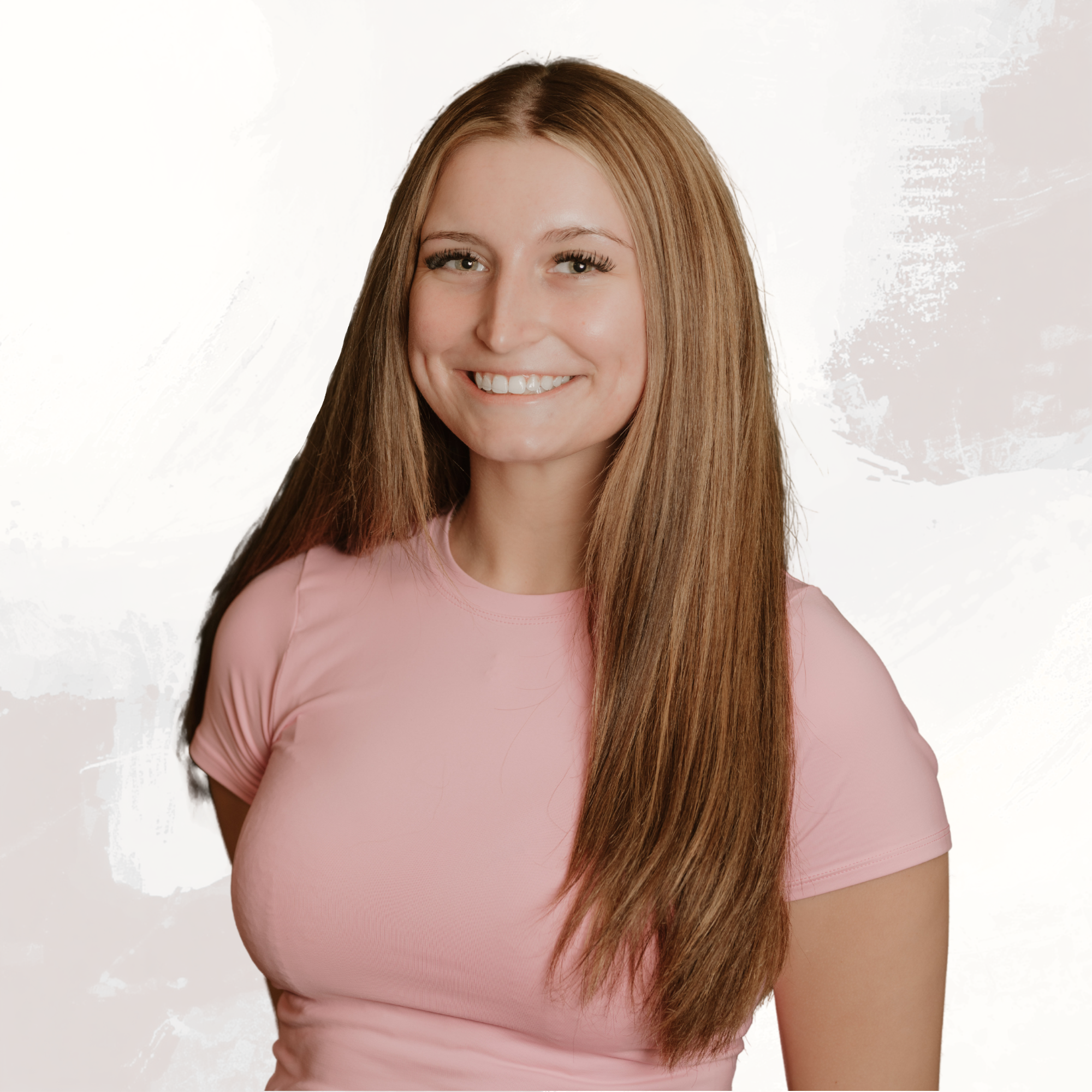 Close-up portrait of a young woman with long brown hair, smiling, wearing a pink top, against a light background.