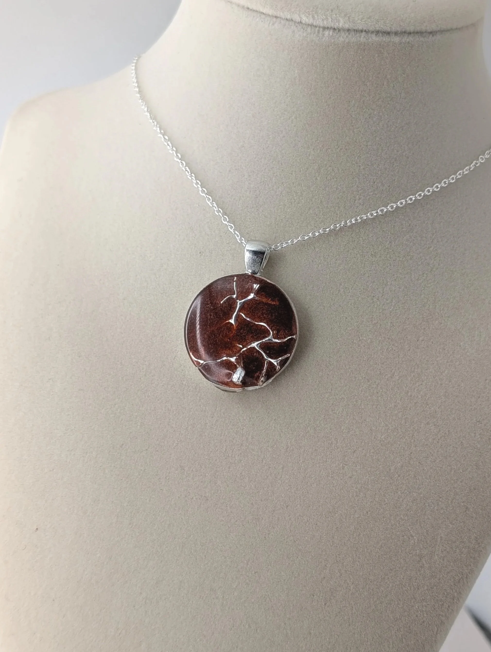 A silver necklace with a circular pendant featuring a tree design on a beige display stand.
