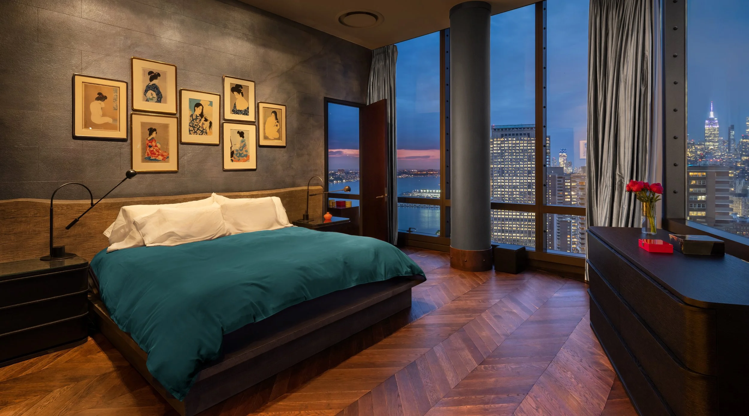 A modern bedroom with a large window showing a city skyline at dusk, featuring tall buildings and a river. The room has a bed with a green duvet and white pillows, framed artwork of women in traditional attire on the wall, a nightstand with a lamp, a