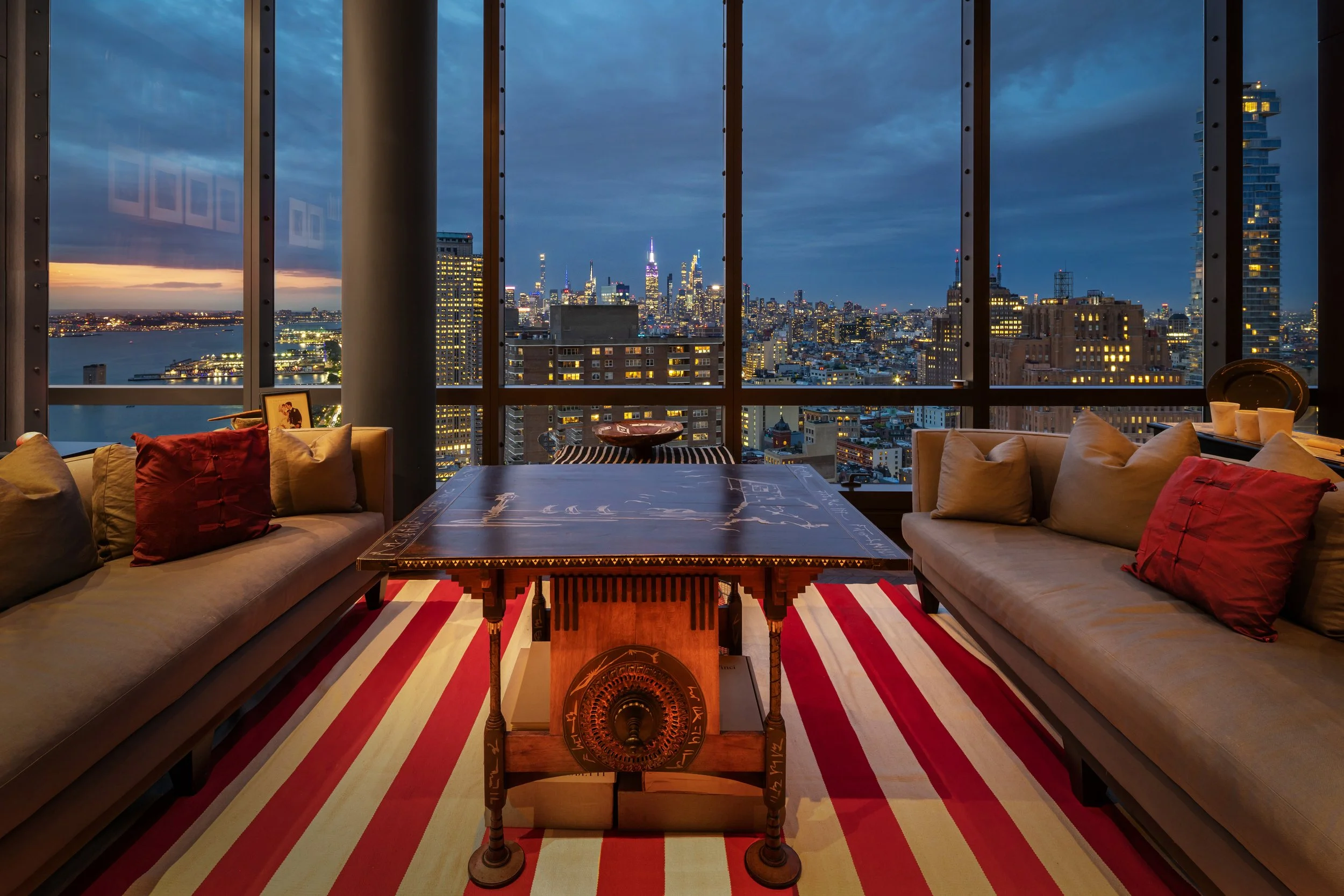 Interior of a high-rise apartment living room at dusk, with large windows showing a city skyline of skyscrapers, a river, and cloudy sky, furnished with sofas, a striped rug, and a wooden table.