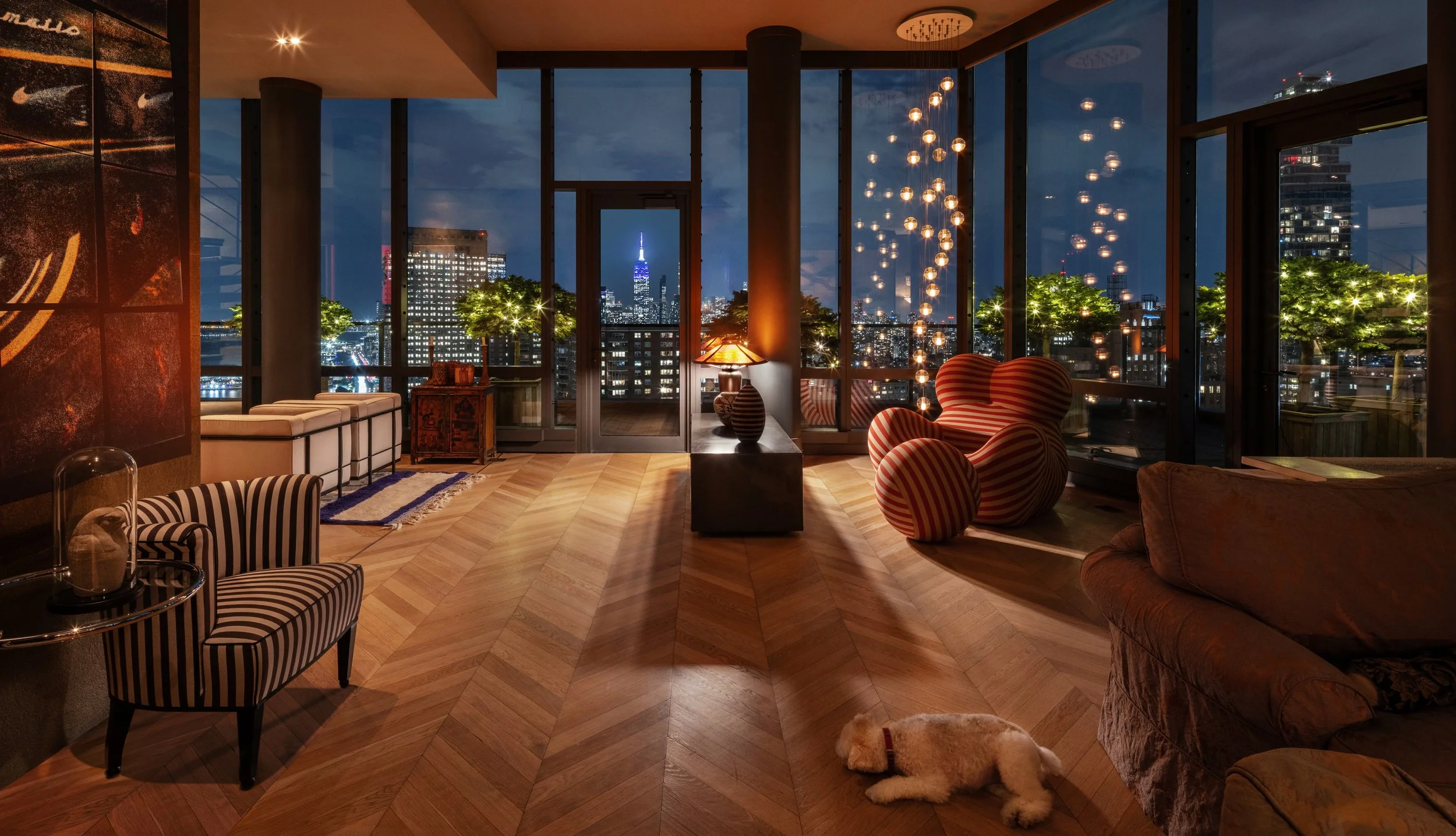 Luxurious living room at night with floor-to-ceiling windows revealing a city skyline with the Empire State Building illuminated. The room features modern furniture, including a striped armchair, a patterned sofa, and a cluster of hanging pendant lig