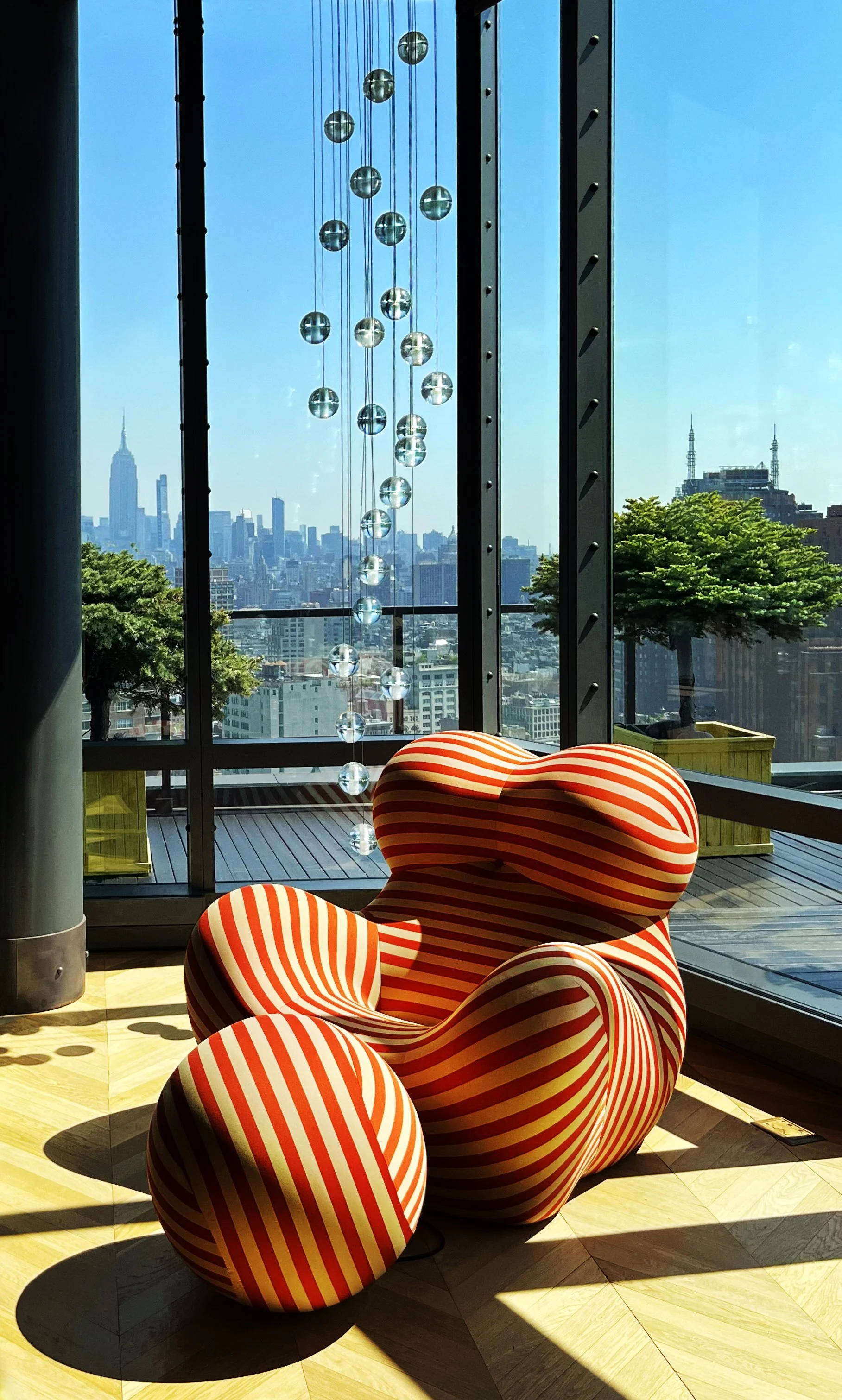 A modern high-rise apartment with a view of New York City skyline, featuring a red and white striped armchair and a glass chandelier with crystal spheres.
