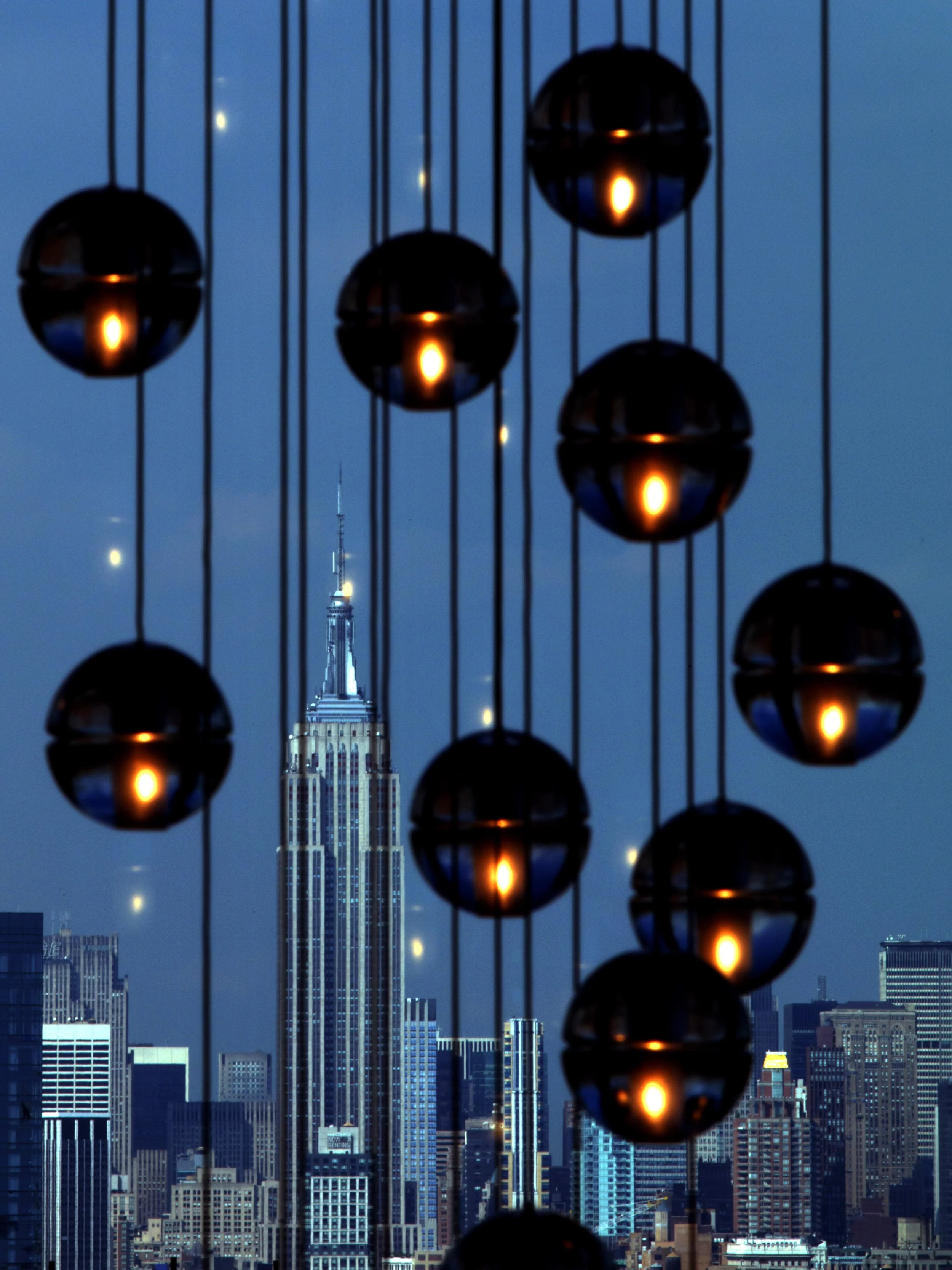 Dusk view of New York City skyline featuring the Empire State Building, framed by a hanging spherical light decoration with warm light reflections.