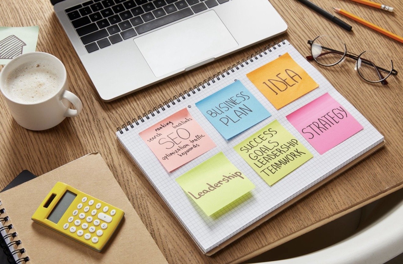 Top view of a desk with a laptop, a cup of coffee, glasses, a yellow calculator, notebooks, and colorful sticky notes with planning ideas.