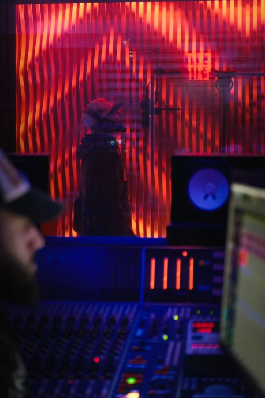 A person wearing a hat in the foreground of a room with audio equipment, with a colorful, wavy, striped red and black light display on the wall behind.