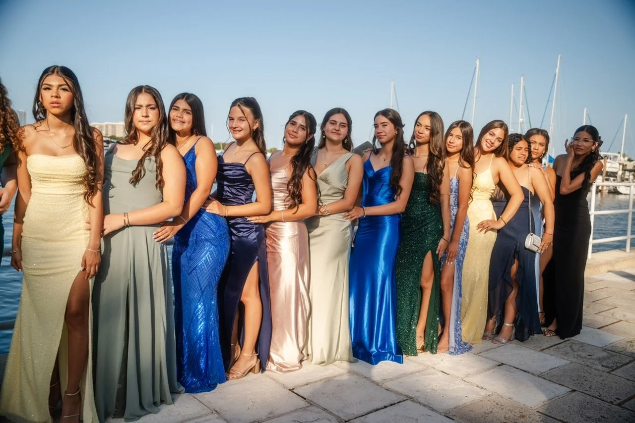 A group of young women in evening gowns standing side by side on a waterfront promenade with boats in the background.