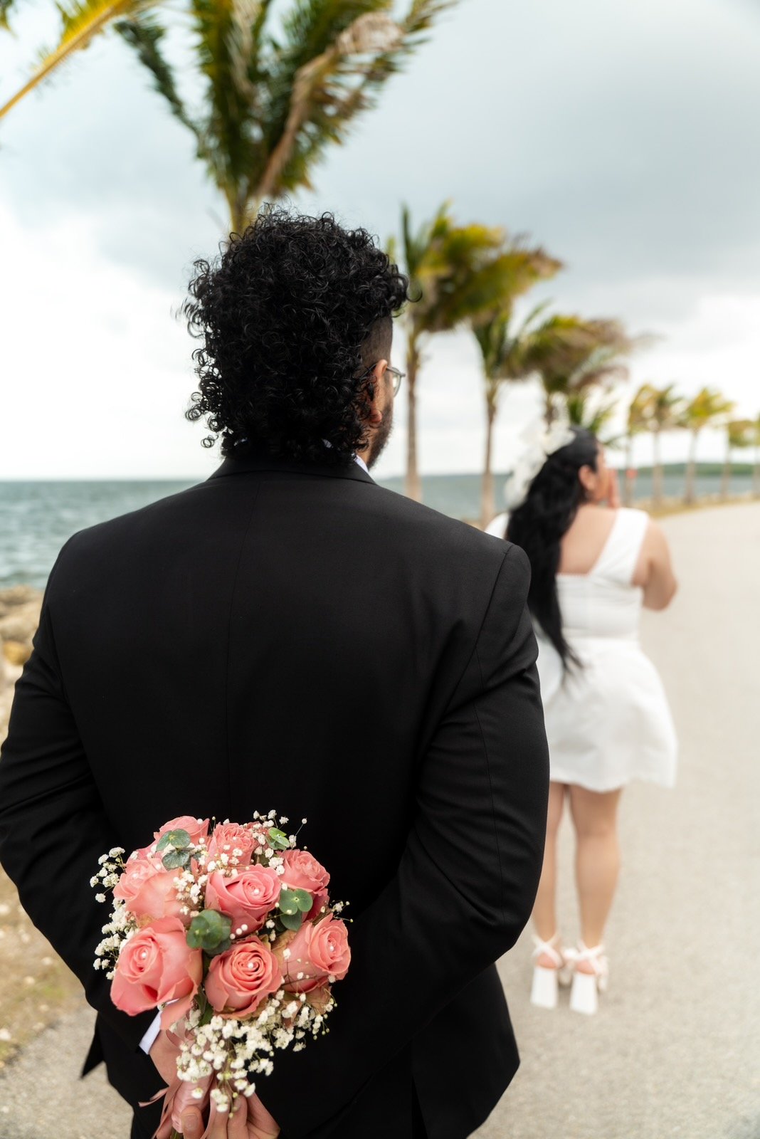 A man in a black suit holding a bouquet of pink roses behind his back, standing on a beachside road with a woman in a white dress walking ahead of him, lined with palm trees and facing toward the ocean.