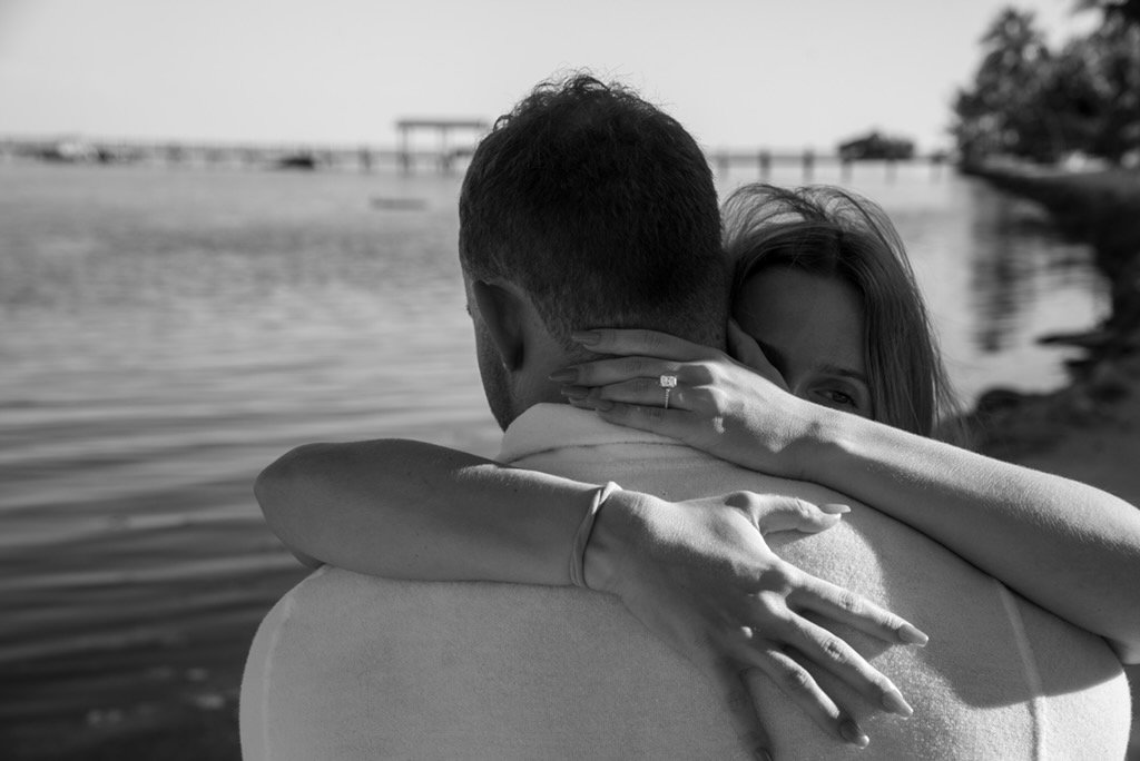 A black-and-white photo of a couple hugging by the water, with a pier in the background.