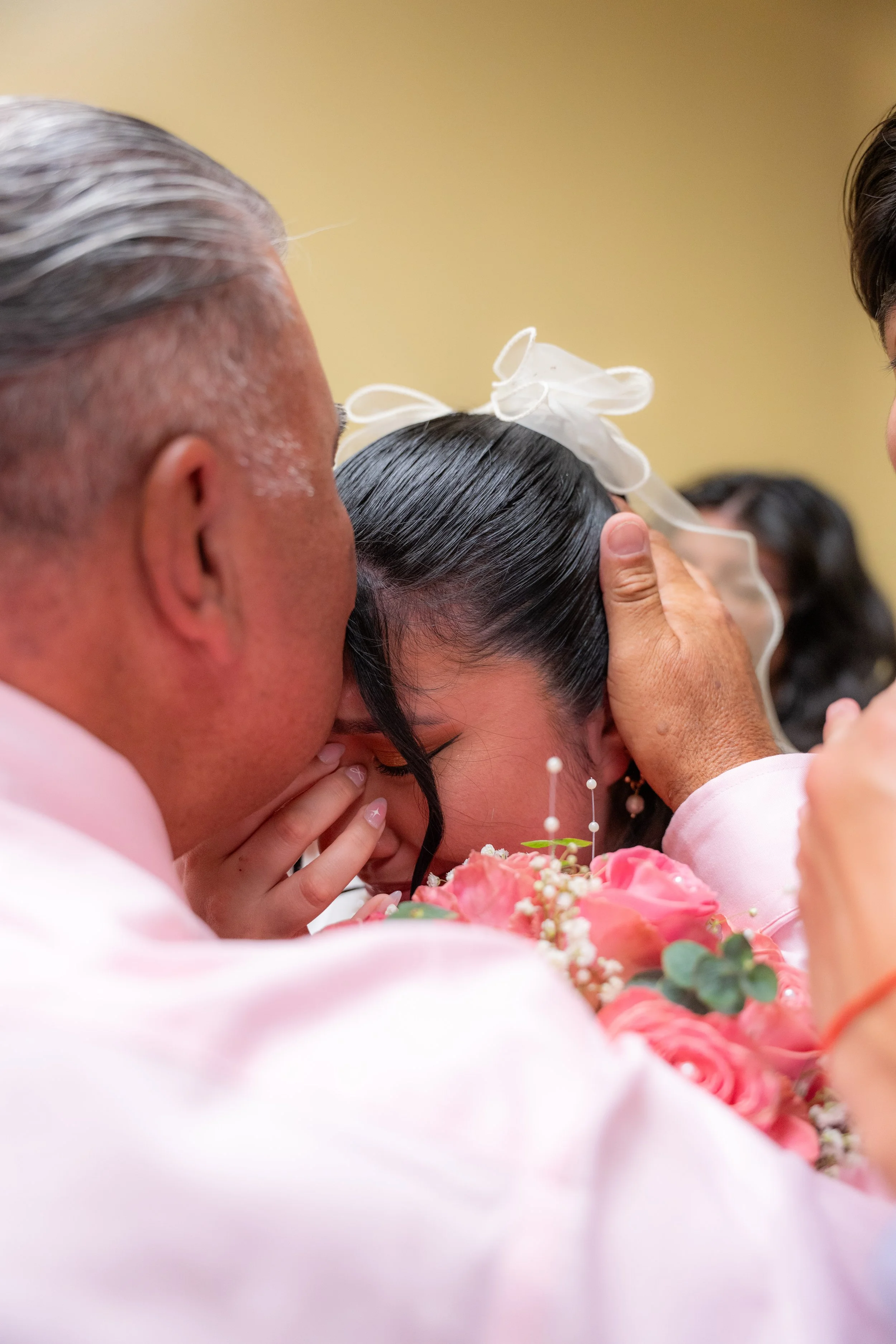 A close-up of a wedding ceremony showing an elderly man, a woman with dark hair and a white bow, and another person in pink holding a bouquet of pink flowers with baby's breath and greenery, with a yellow wall in the background.