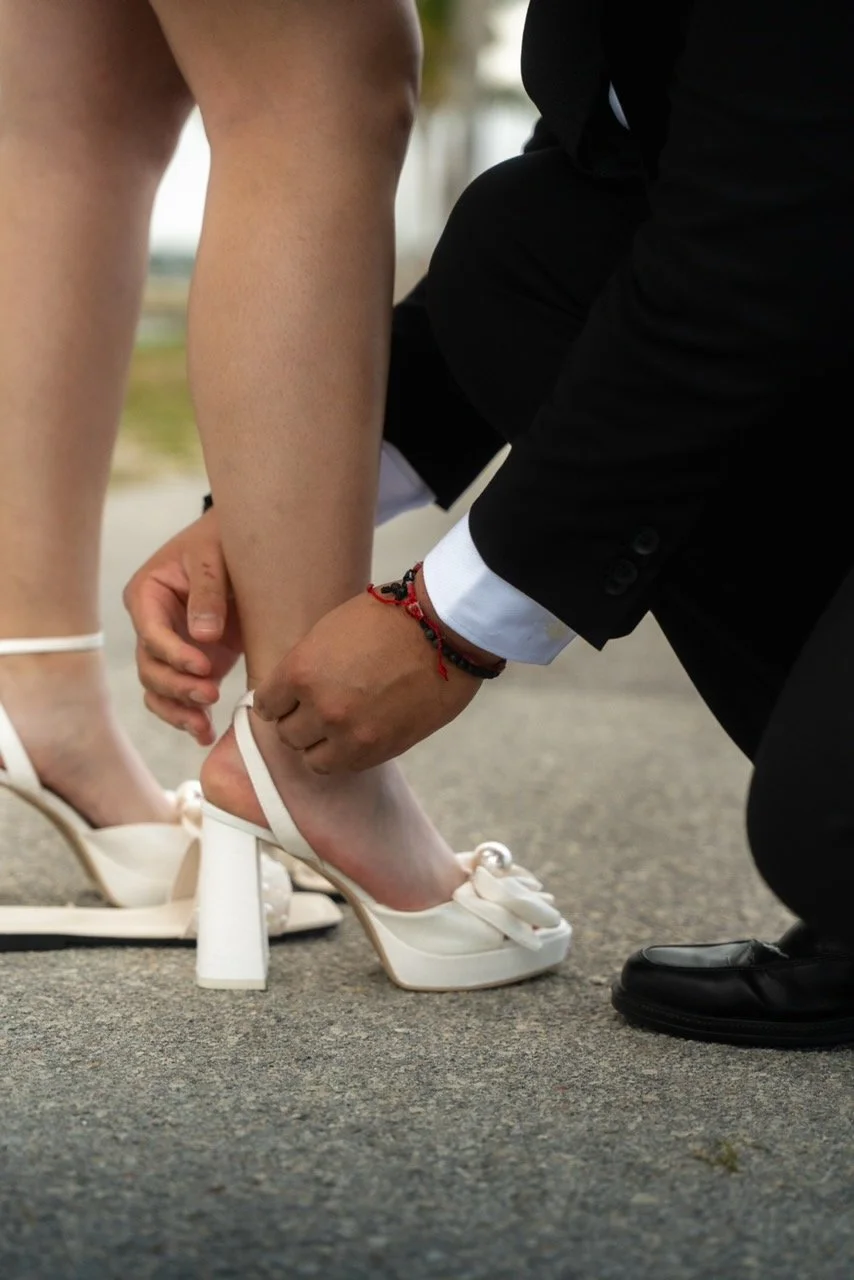 A man in a black suit kneeling and fastening the strap of a woman's white high-heeled sandal, likely during a wedding or formal event.