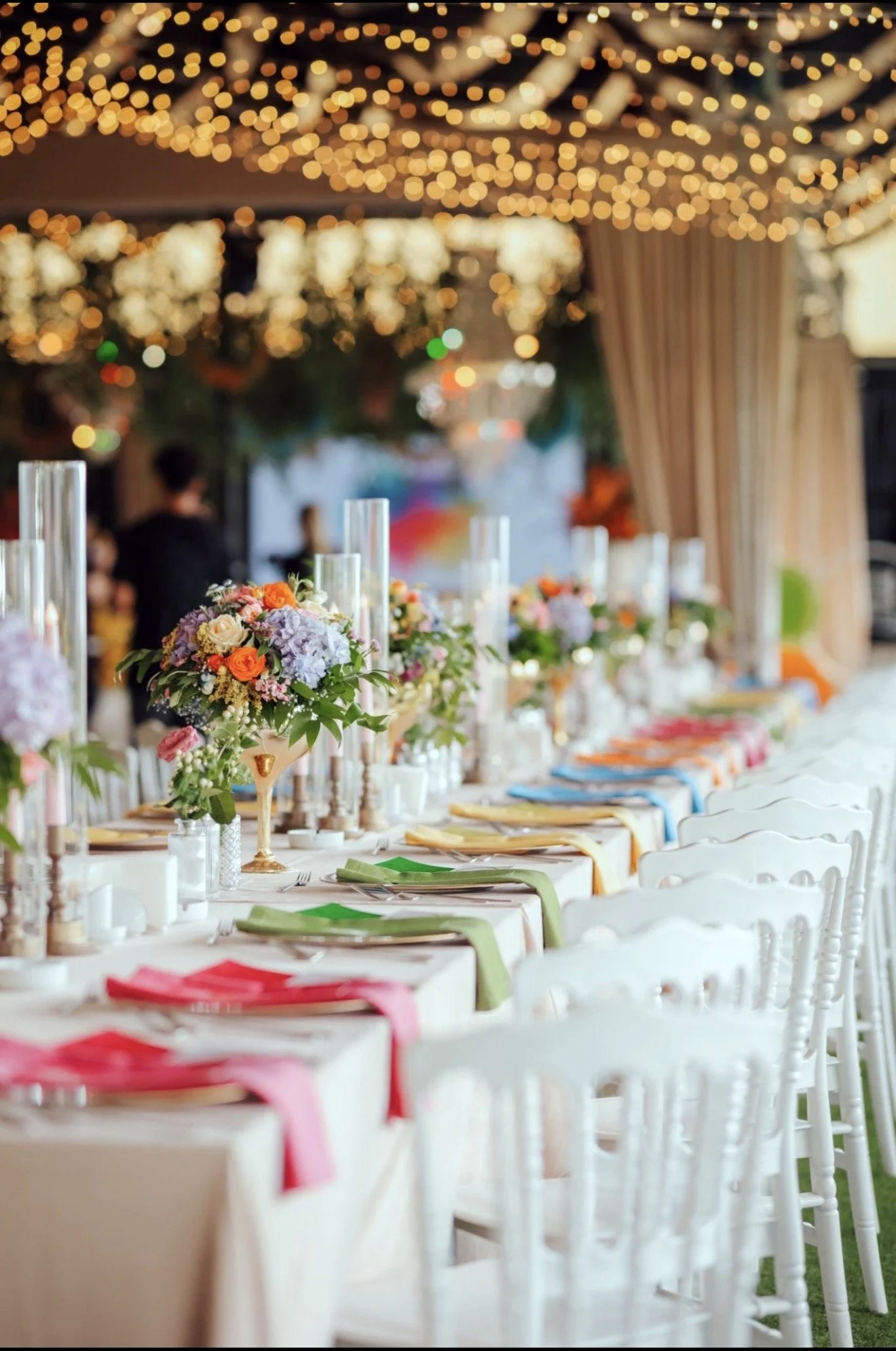 A decorated banquet table at an event venue with floral centerpieces and colorful napkins, with string lights hanging from the ceiling, creating a festive atmosphere.