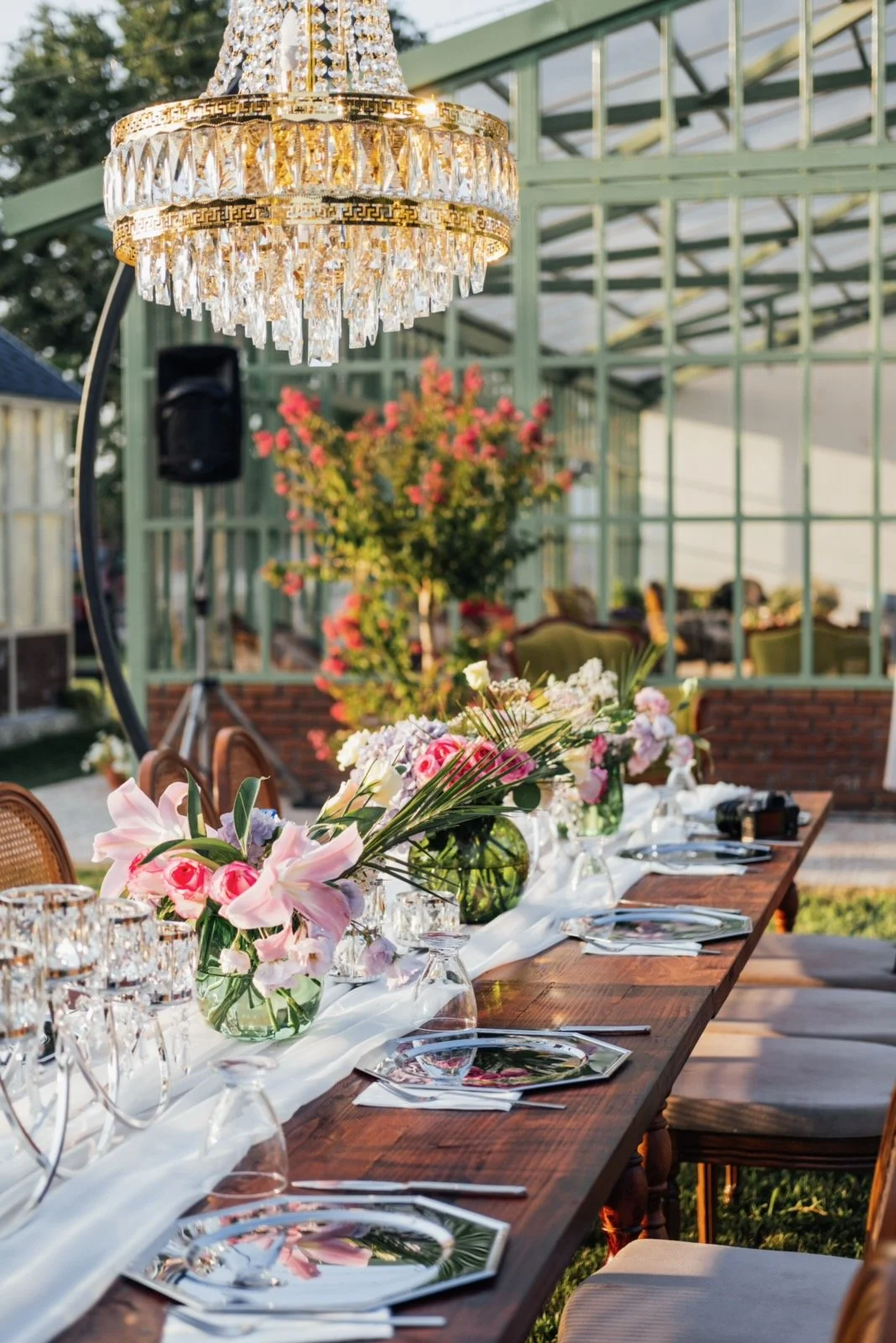 A long wooden table set for a meal outdoors under a large chandelier, with floral centerpieces, glassware, plates, and napkins, in a garden setting with plants and a greenhouse-like structure in the background.
