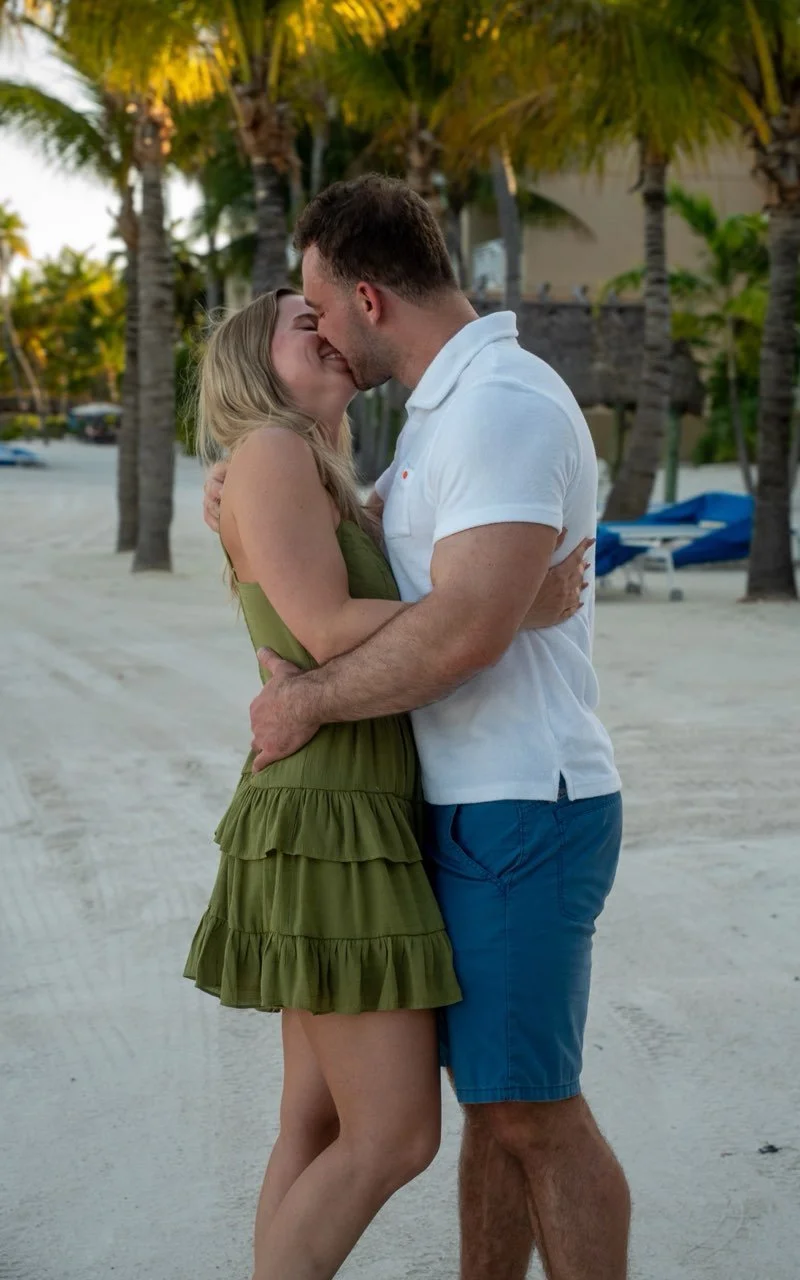 A couple kissing on the beach, with palm trees and boats in the background.