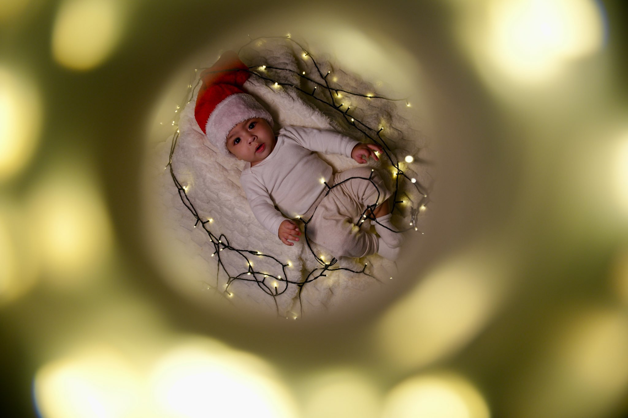 Baby wearing a Santa hat lying on a blanket surrounded by string lights, viewed through a circular frame.