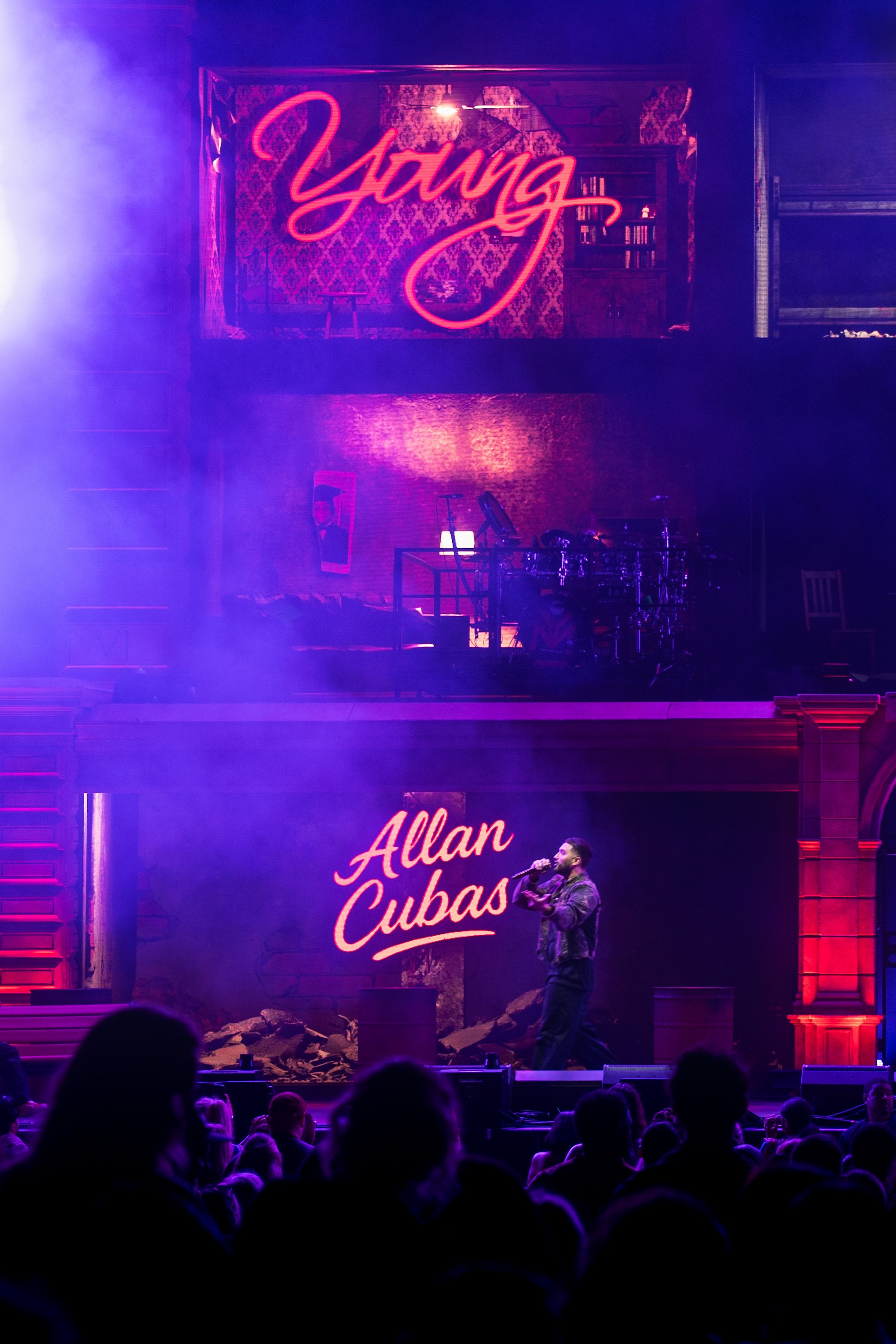 A man is performing on stage with a microphone, in front of a large sign that reads 'Allan Cubas'. The stage has purple lighting, a brick wall backdrop, and a second level with a neon sign that says 'Yanna'. There are audience members visible in the foreground.