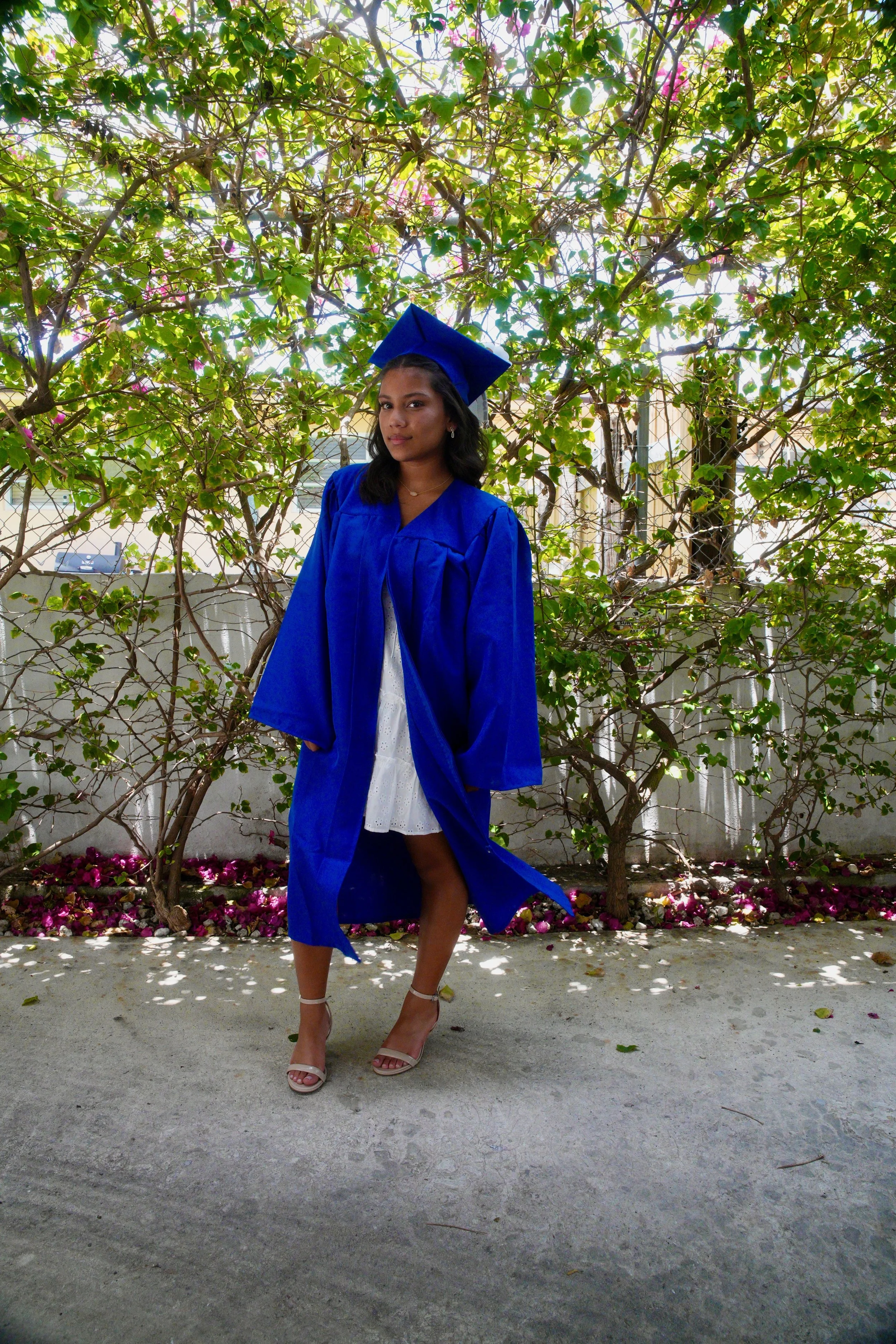 A young woman wearing a blue graduation gown and cap stands outdoors in front of a leafy green hedge with pink flowers. She is looking at the camera with a neutral expression, wearing white open-toe heels and a white dress.