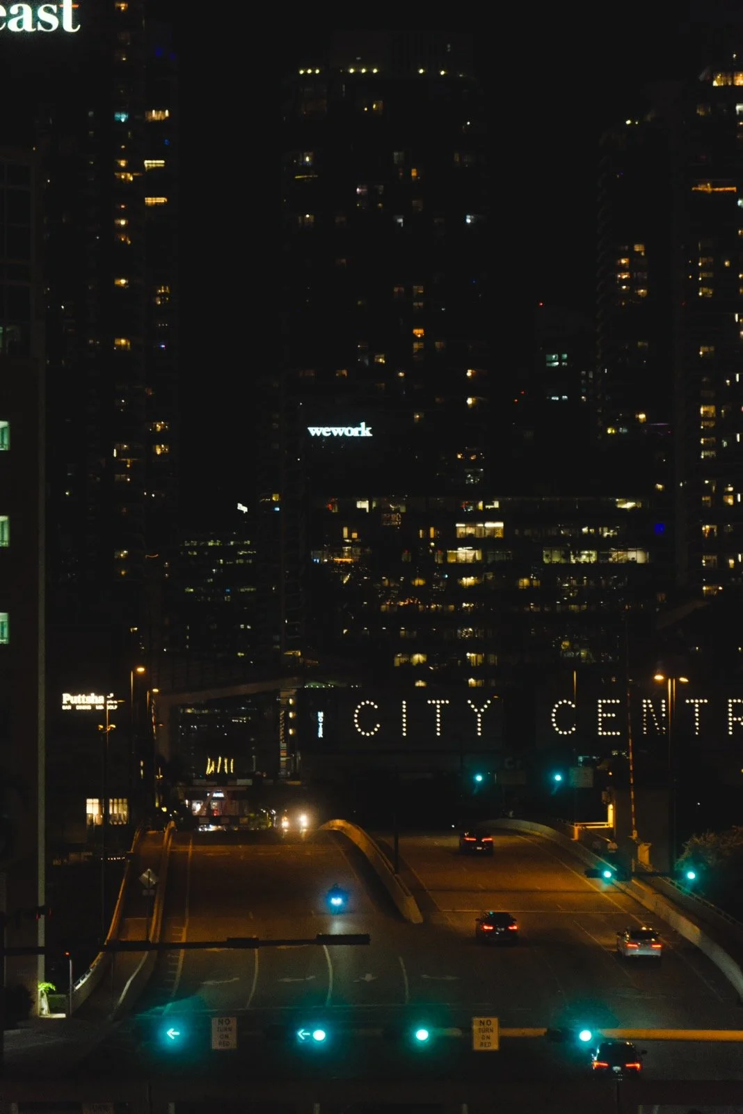Nighttime cityscape with illuminated high-rise buildings, visible signs reading 'wework' and 'City Center,' and a few cars on the road.