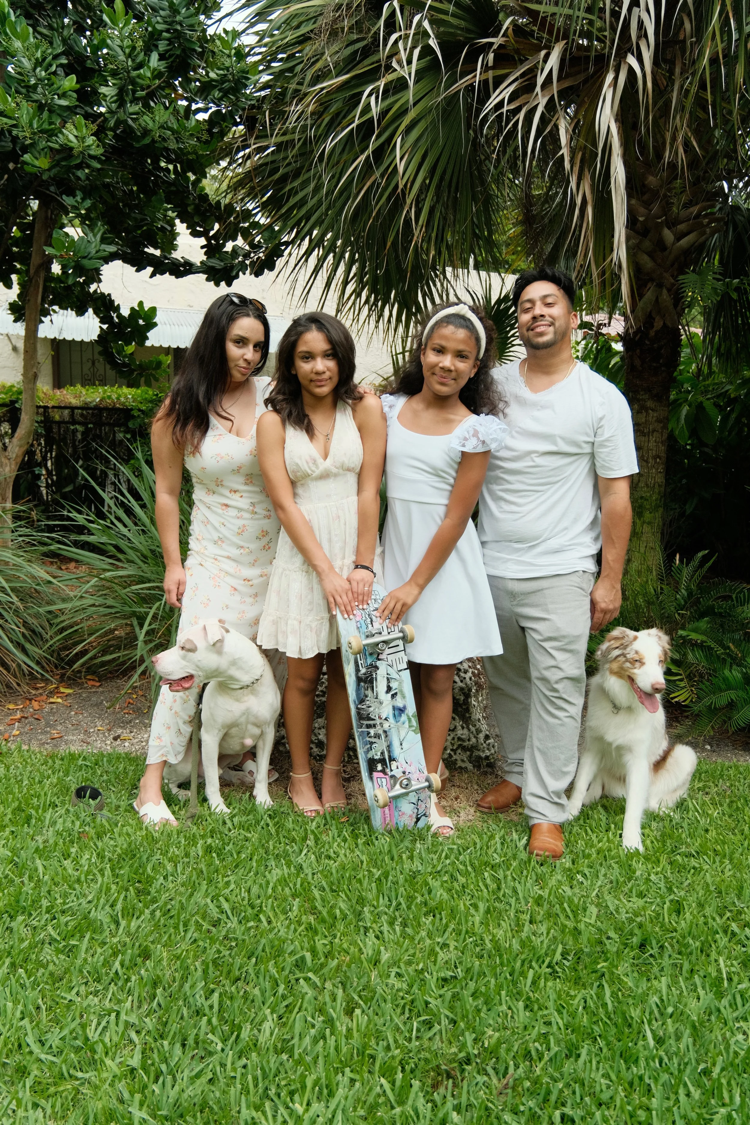 Four young people standing outdoors in a garden with two dogs, one of which is a white Pitbull, and a skateboard, surrounded by lush green plants and trees.