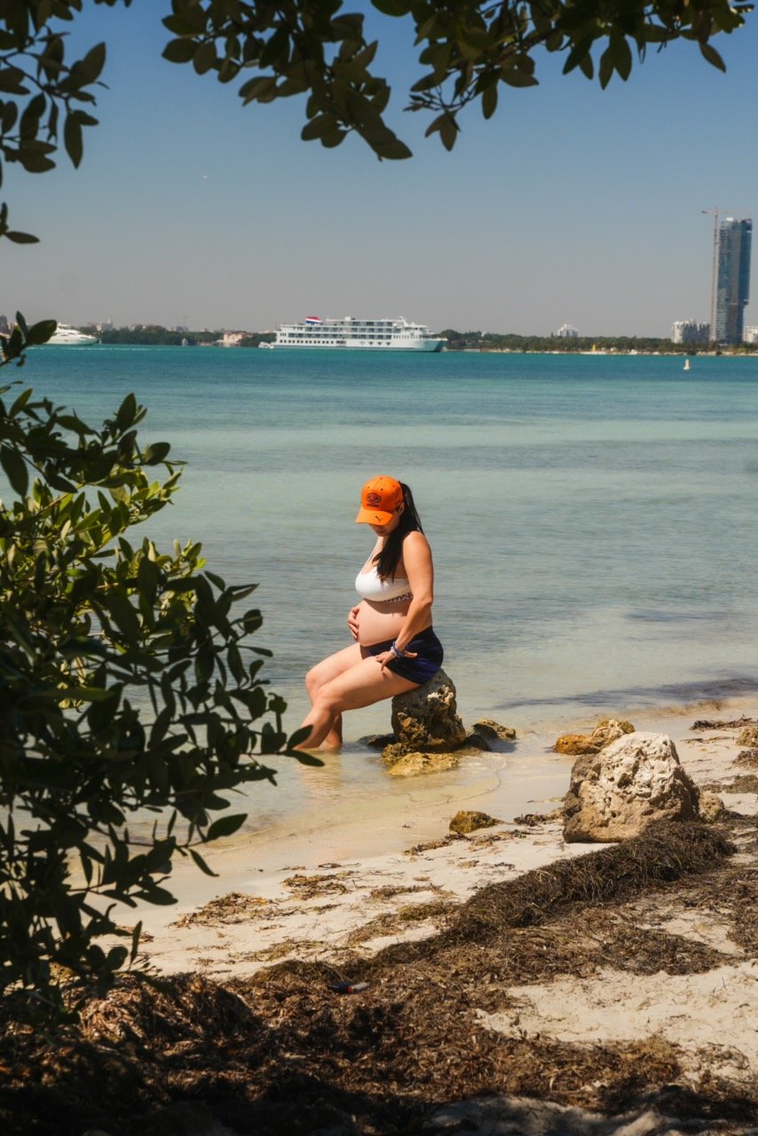 A pregnant woman sitting on rocks at the edge of a beach, looking down, with water and boats in the background, partially shaded by trees and greenery.