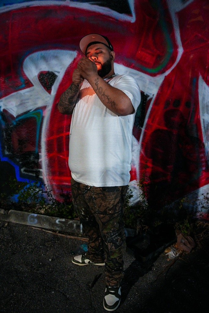 A bearded man in a white t-shirt, camouflage pants, and a cap stands in front of a colorful graffiti wall, with his hands clasped together near his face.